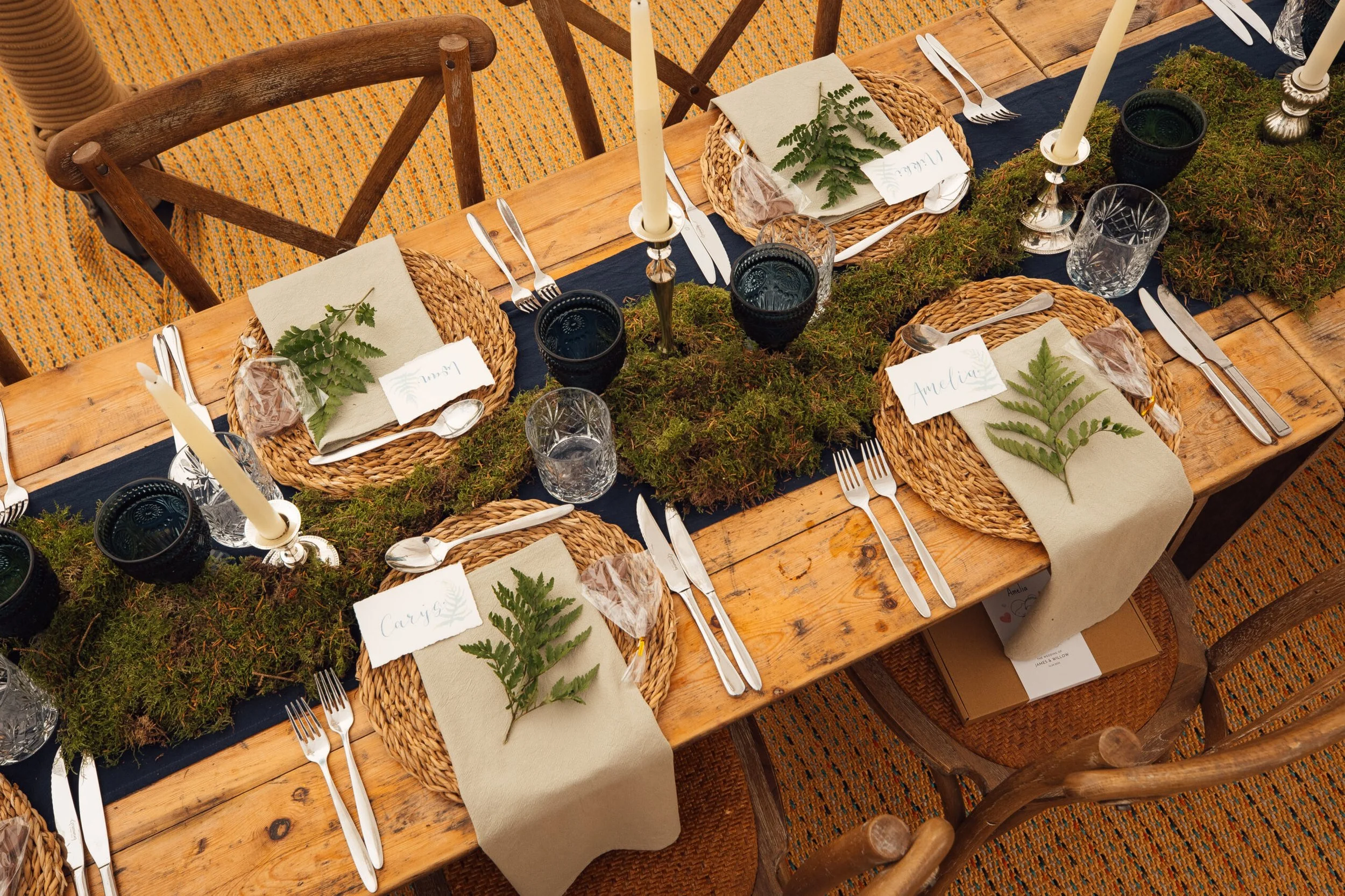 A wooden dining table set for a meal with woven placemats, napkins with fern leaves, silverware, black and clear glasses, and white candles with gold holders. The table is decorated with moss and greenery.