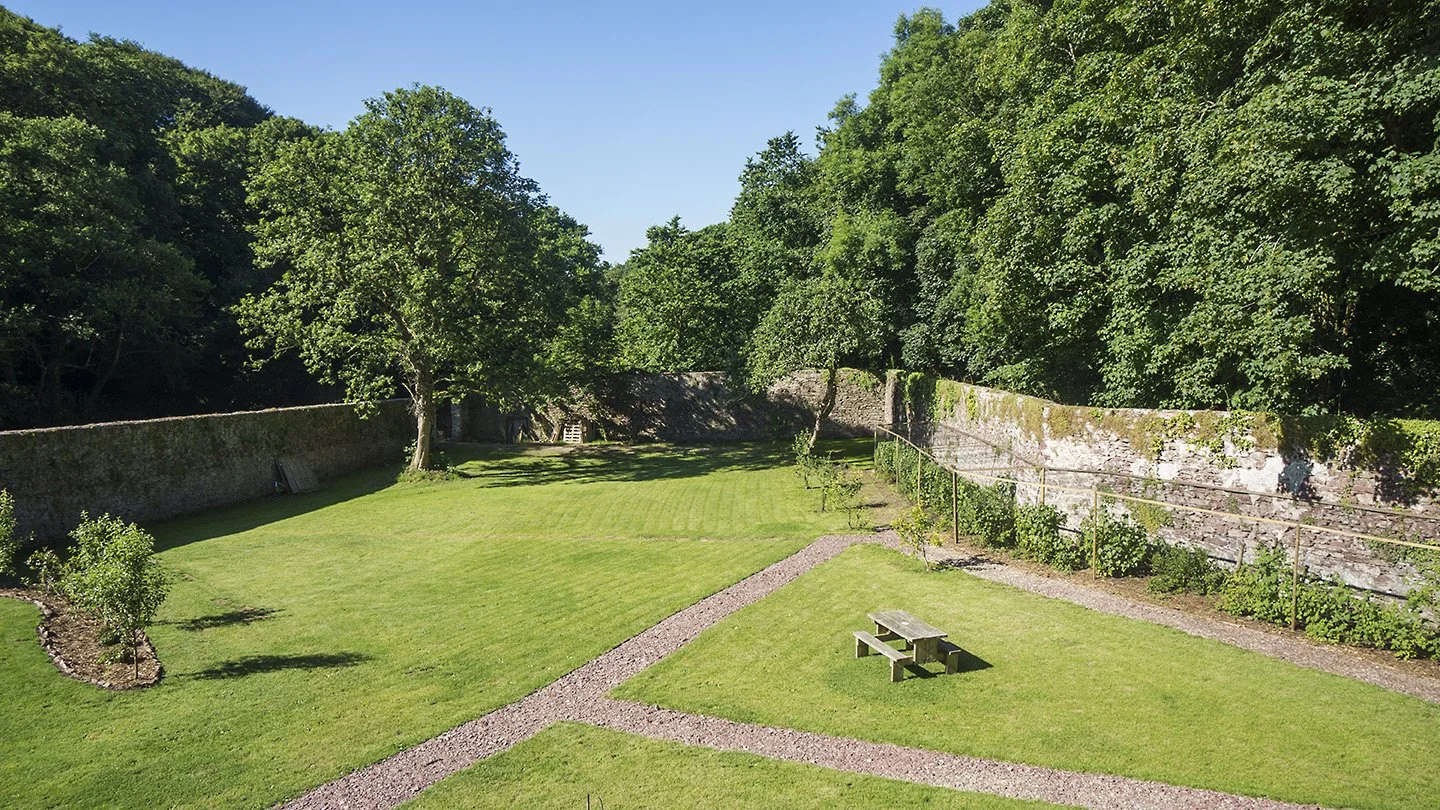 A lush green park with a small picnic table, surrounded by a stone wall and tall trees with dense foliage