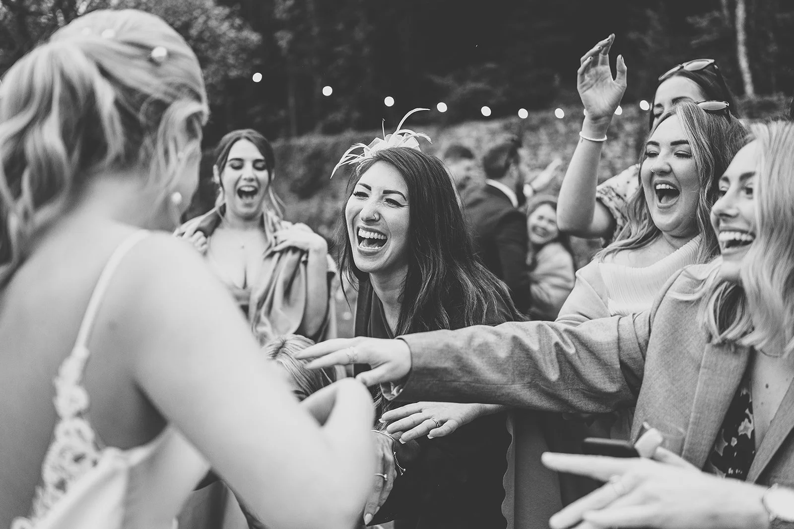 Black and white photo of a group of women celebrating outdoors, laughing and smiling, dressed casually, with string lights above in a natural setting.