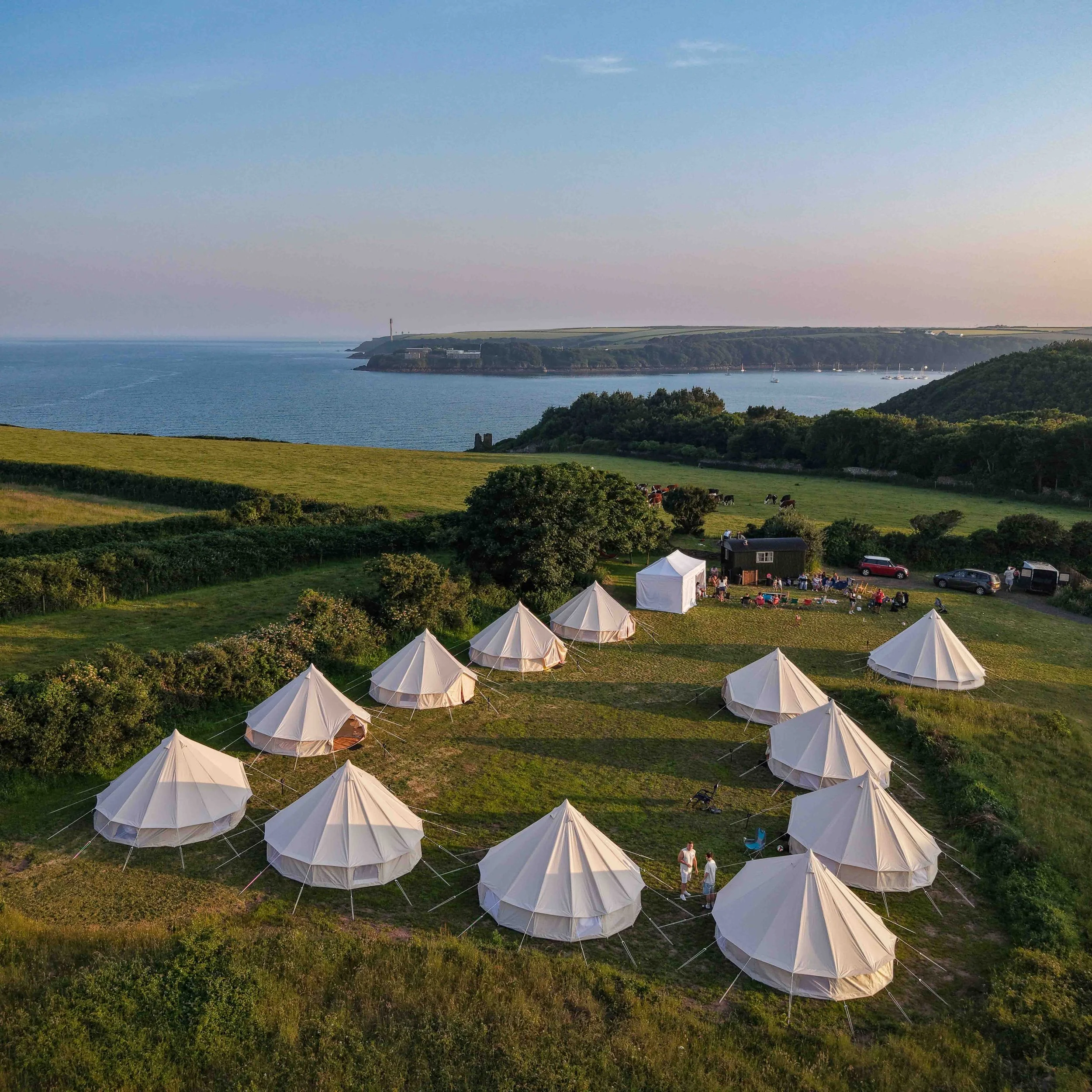 Open field with white tents set up for an outdoor event, overlooking a body of water with boats and a distant coastline, during daytime with clear sky.