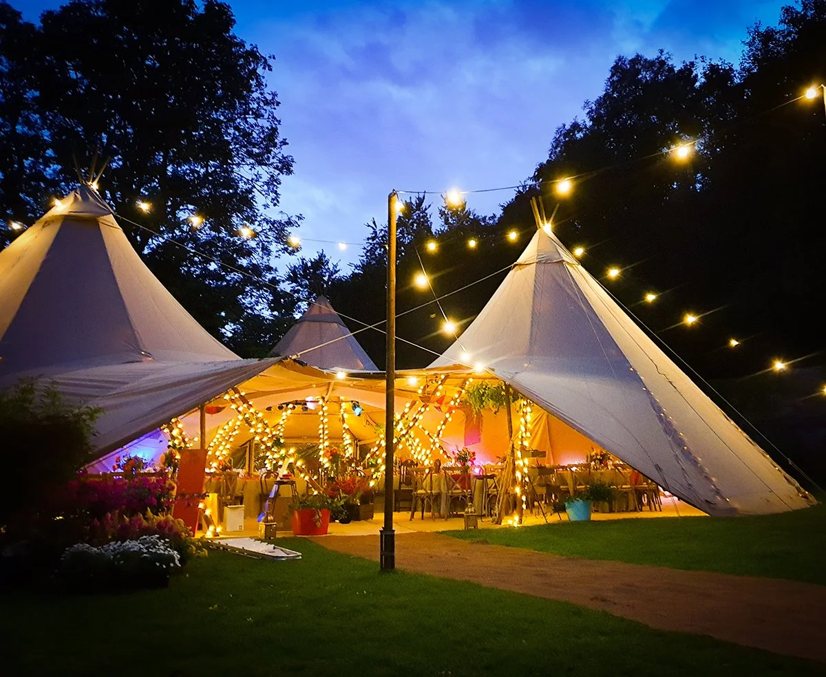 An outdoor event tent with pointed peaks, decorated with string lights, set up in a garden area during evening or dusk, with trees in the background and some potted plants around.