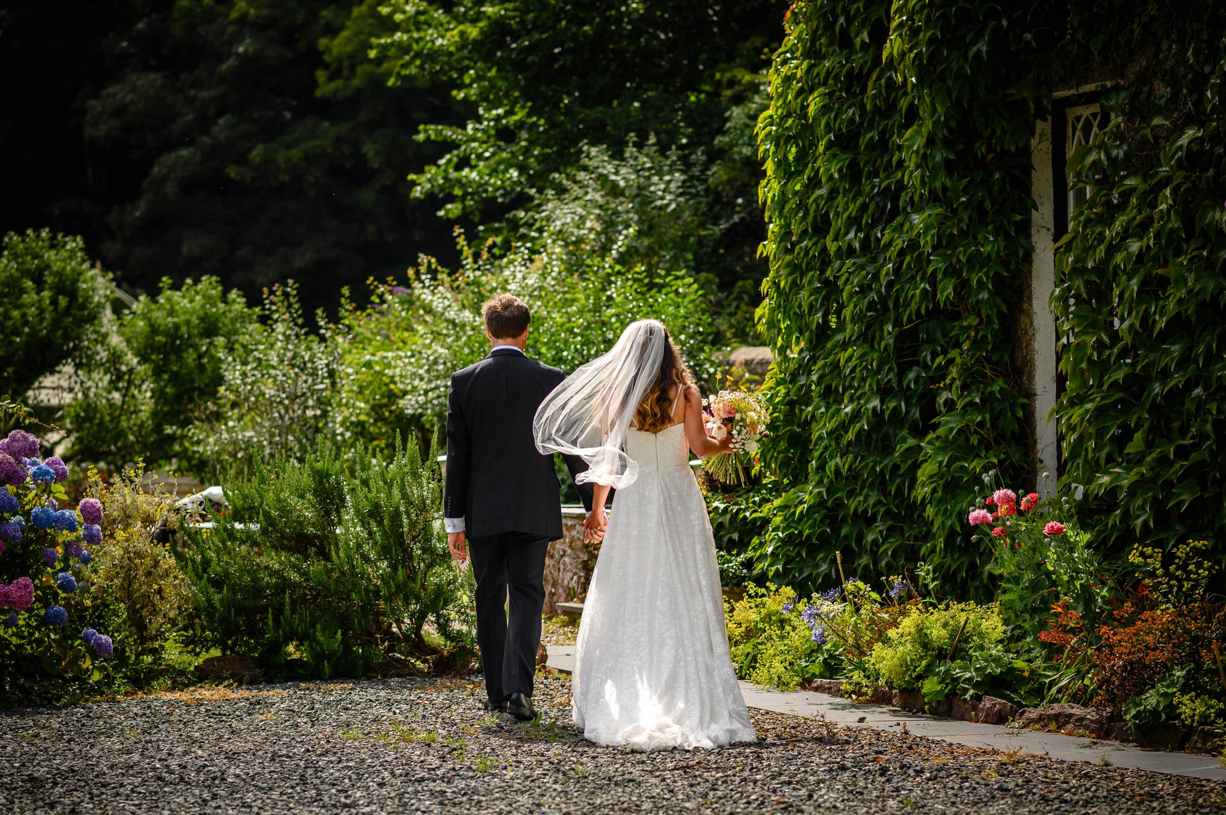 A bride and groom holding hands while walking through a lush garden path surrounded by colorful flowers and greenery, with the bride wearing a white lace wedding gown and veil, and the groom dressed in a black suit.