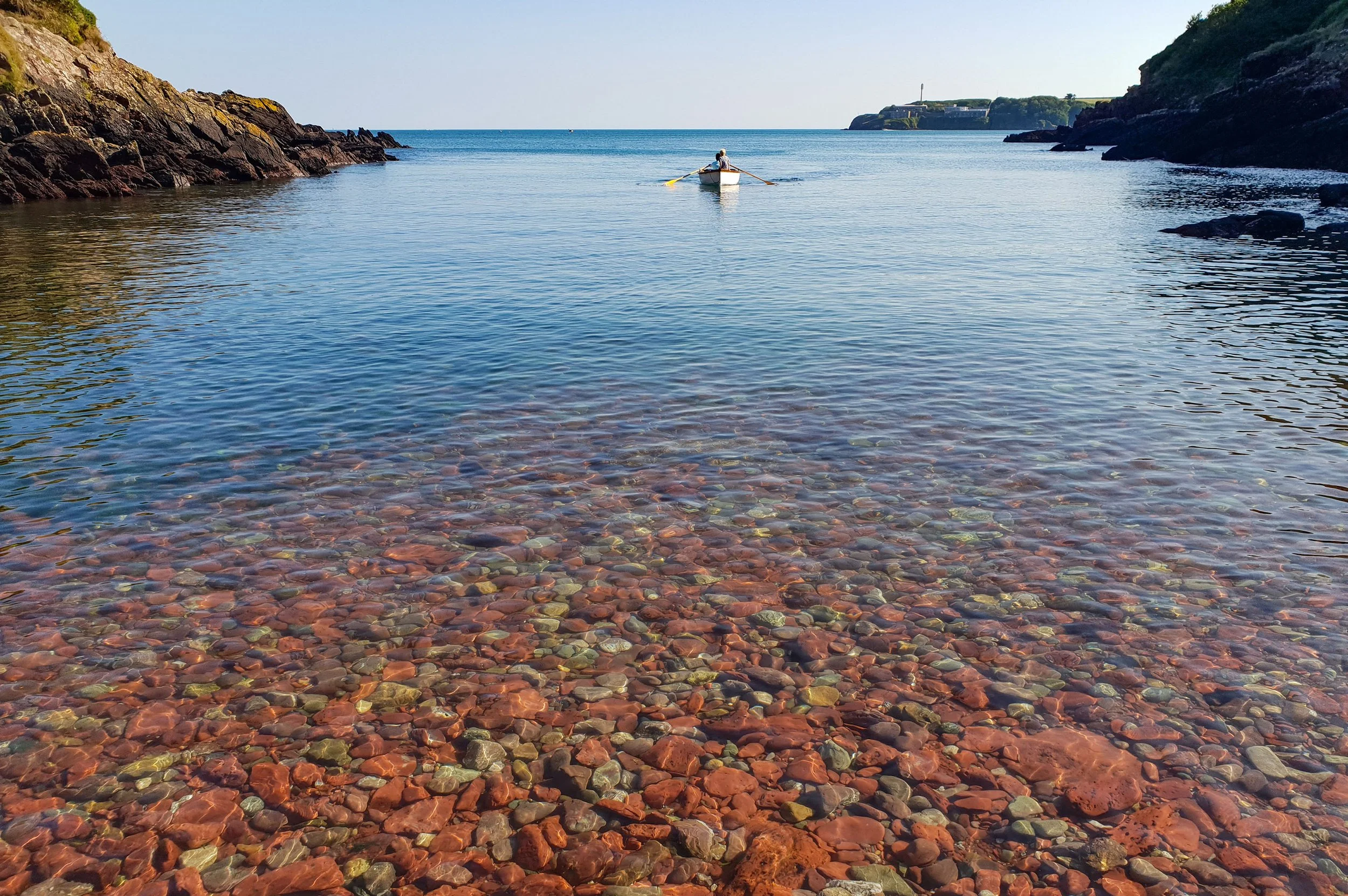 A person rowing a small boat in calm water between rocky cliffs on either side. The water is clear, revealing colorful pebbles at the bottom, and there is a distant shoreline with a lighthouse on a hill.