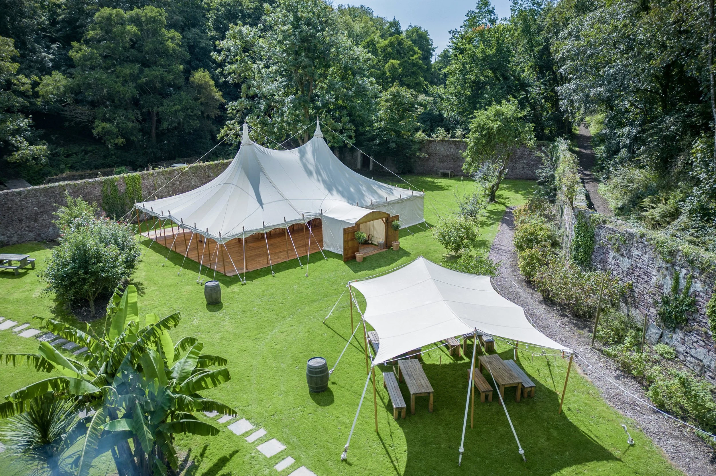 A large white event tent set up on a grassy area surrounded by trees and stone walls, with smaller white canopies and picnic tables nearby in a lush outdoor setting.