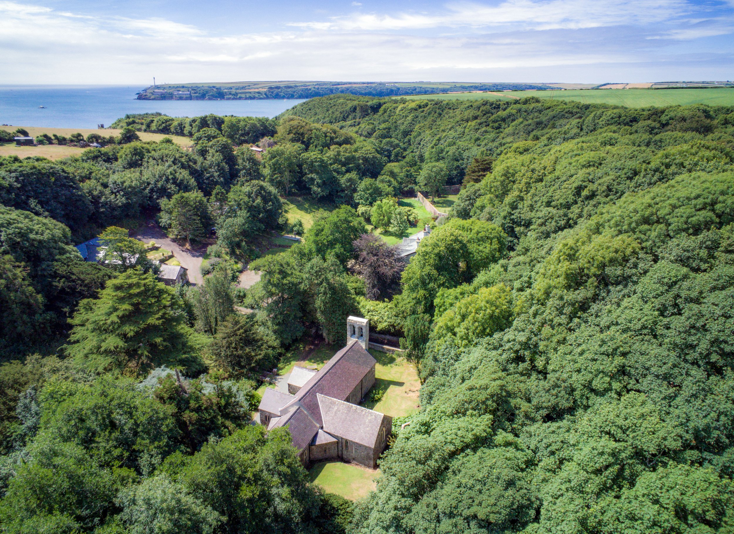 Aerial view of lush green trees surrounding a small cluster of residential buildings with gray roofs, with a body of water and land in the distance under a partly cloudy sky.