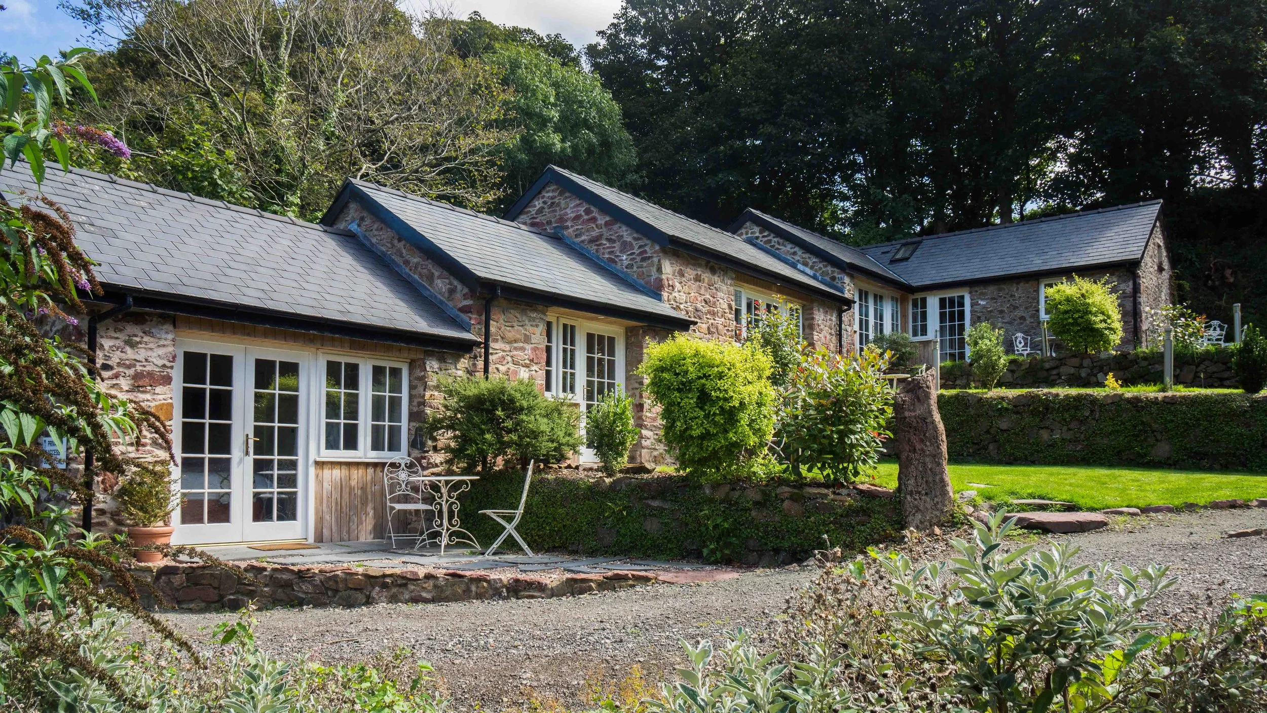 A stone house with a slate roof, surrounded by trees and green bushes, with a patio featuring white chairs and a small table in front.