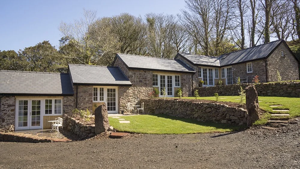 A stone house with multiple sections, gray shingle roofs, white framed windows and doors, a well-kept lawn, stone retaining walls, and outdoor furniture, surrounded by trees and a clear blue sky.
