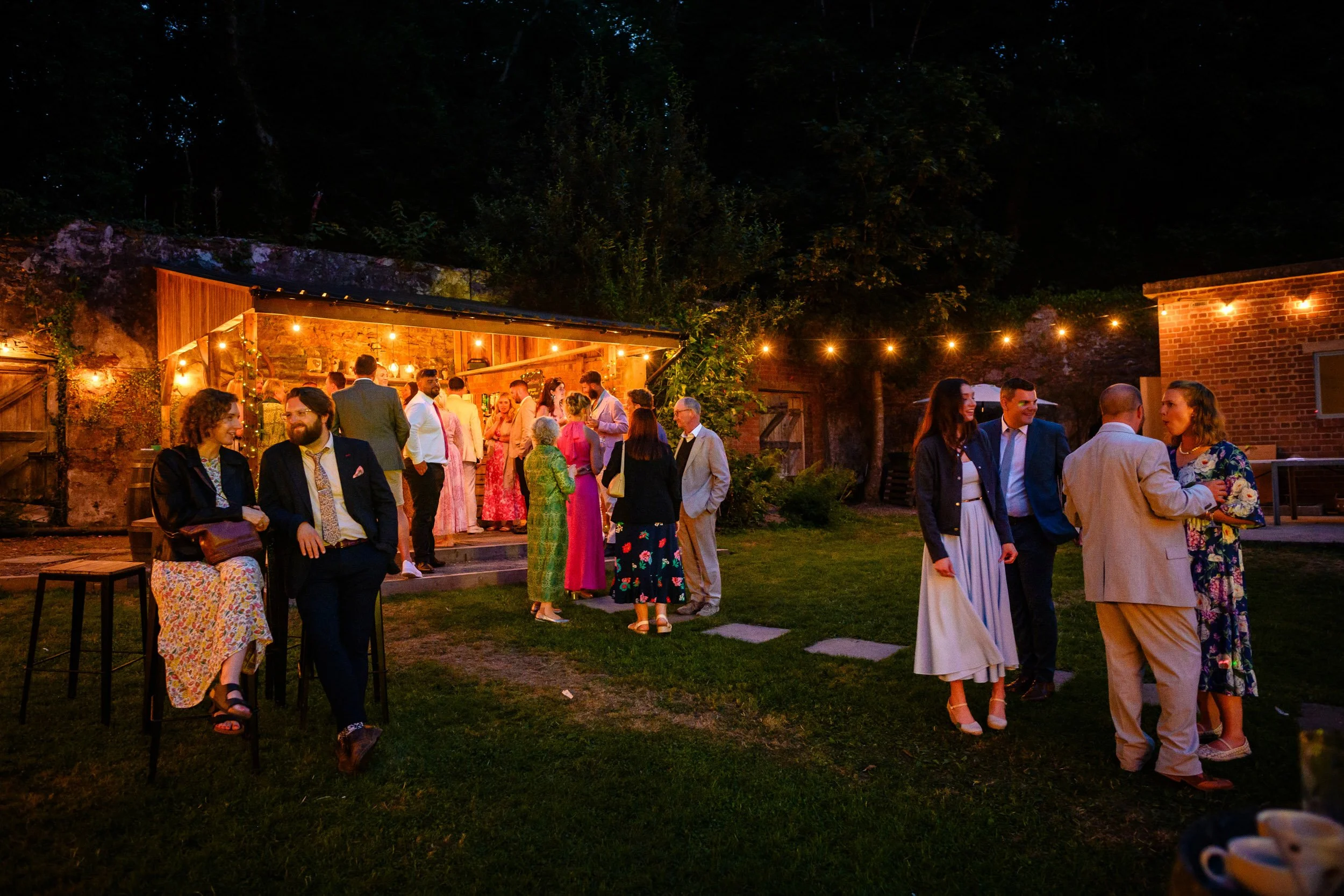 People gathered for an outdoor evening social event with string lights, some standing and some sitting, in a garden with a brick wall and trees in the background.