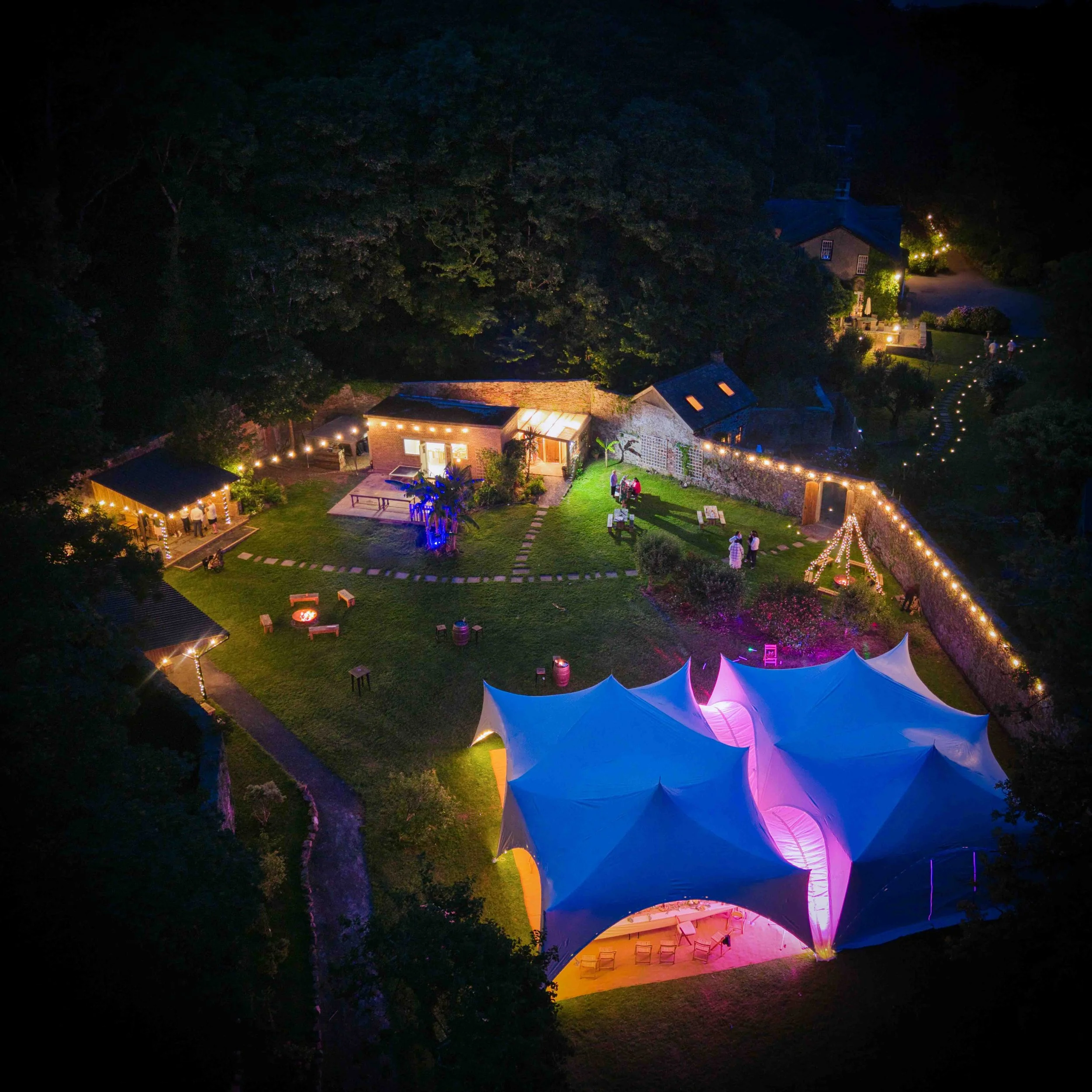 Night view of a backyard with string lights, trees, a house with lit windows, and a tent with colorful lighting, with people gathered outside.