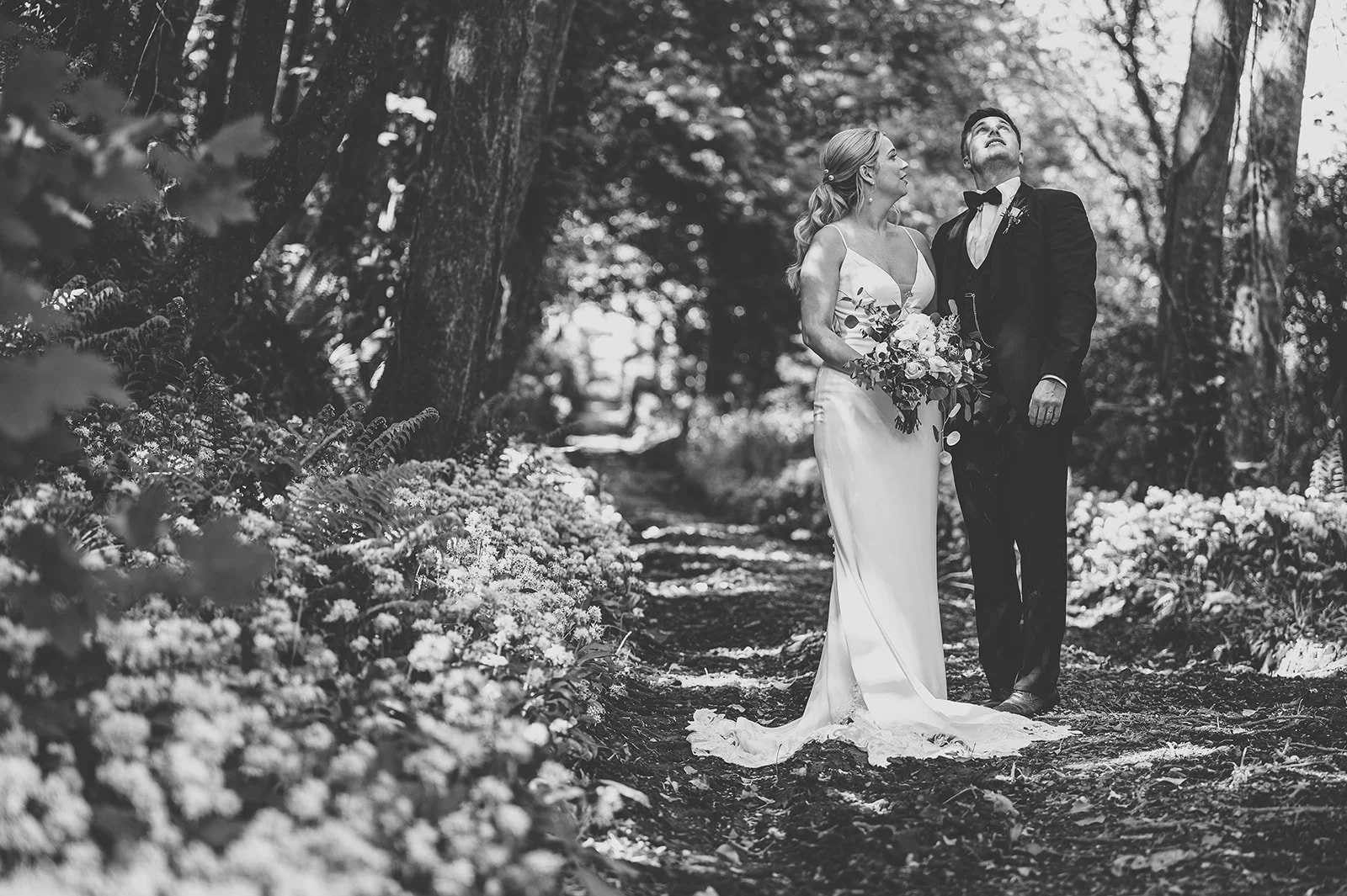Black and white photo of a bride and groom standing on a woodland path, surrounded by trees and flowers, gazing upward.