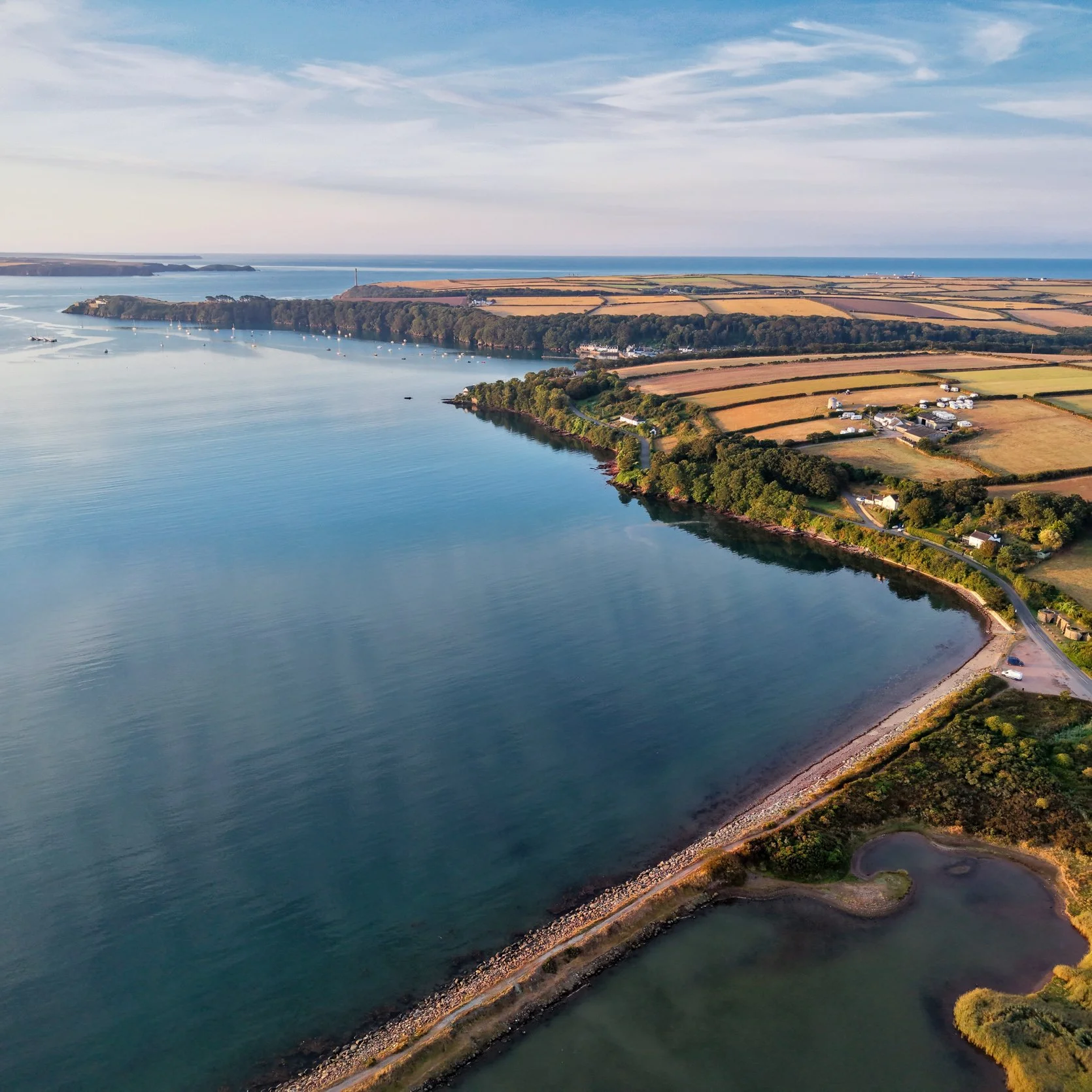 Aerial view of a coastal landscape with a large river estuary, sandy and grassy fields, and small buildings along the shoreline under a blue sky with some clouds.