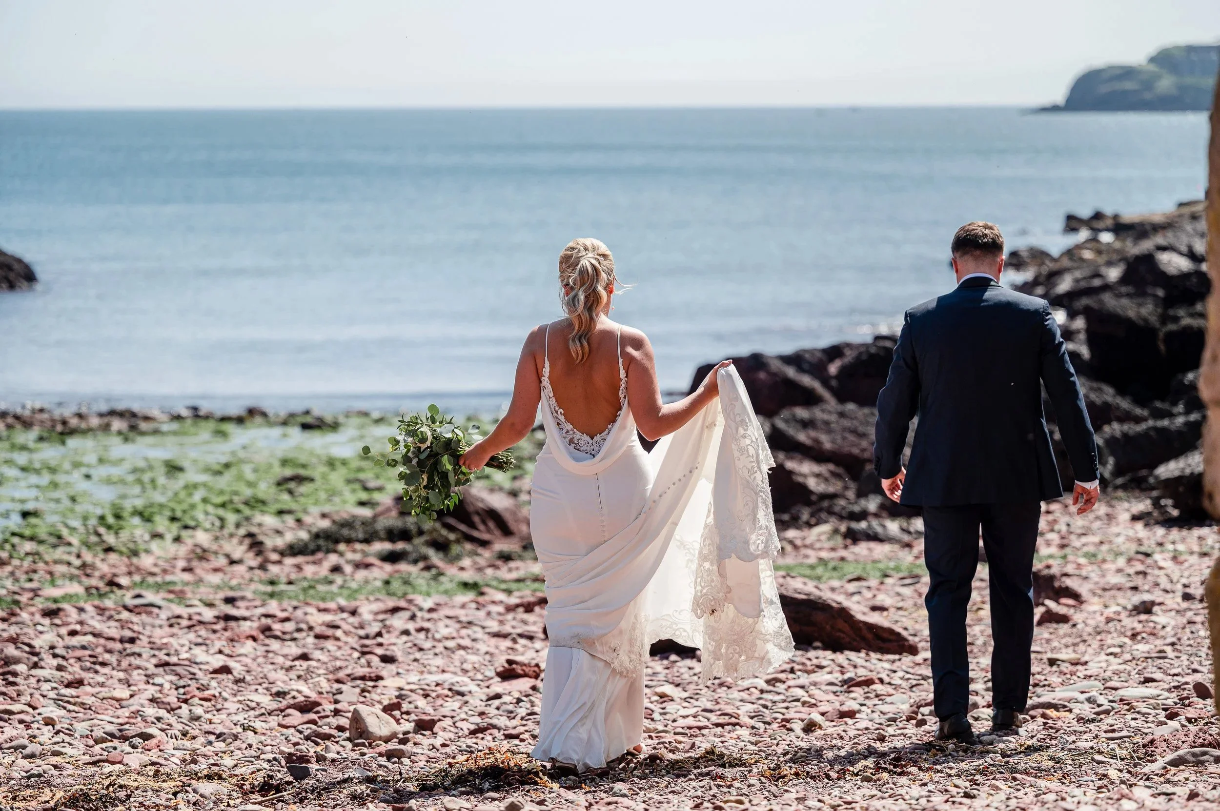 A newlywed couple walking on a rocky beach by the ocean, with the bride in a white wedding gown holding a bouquet and the groom in a dark suit.
