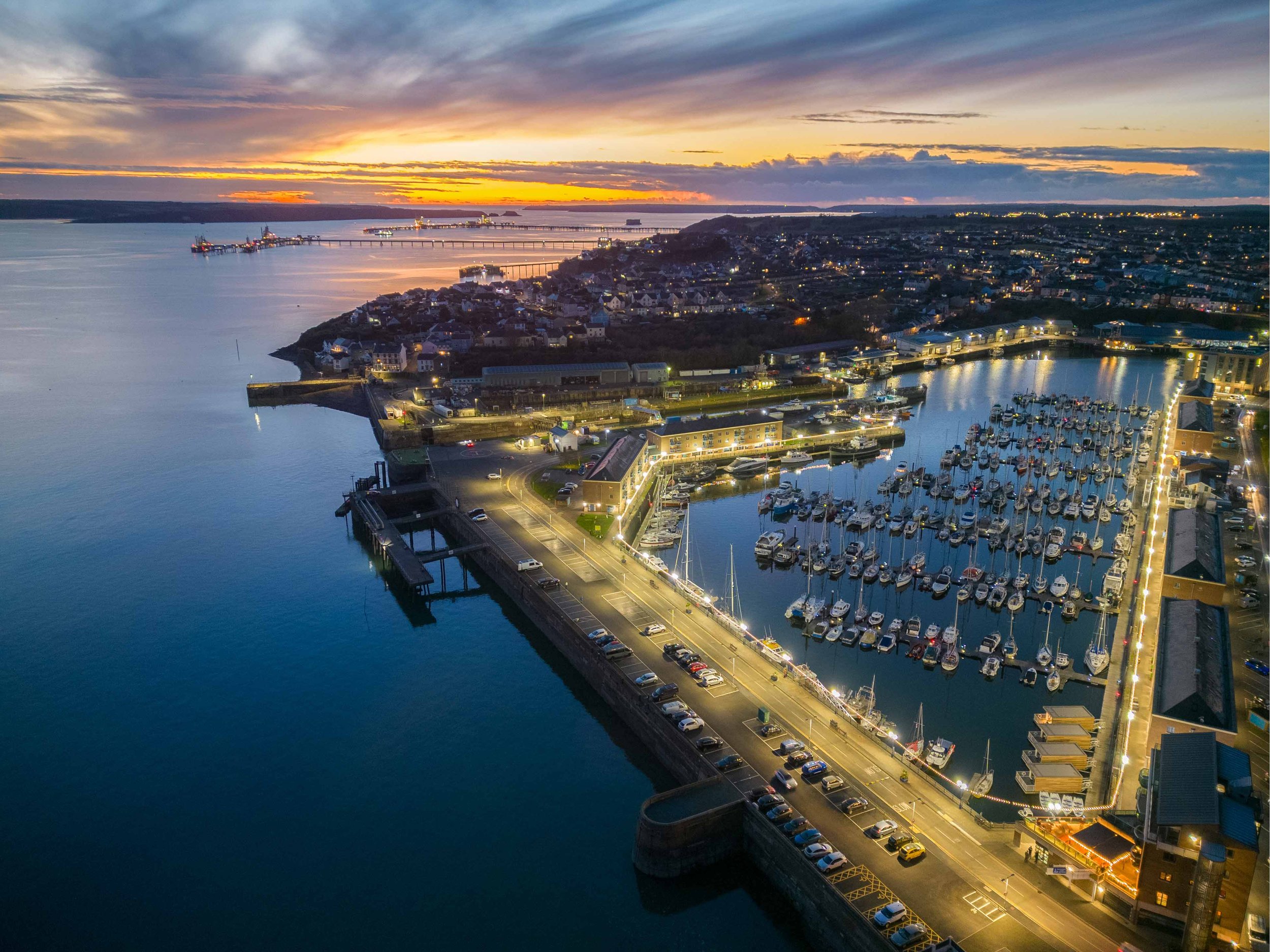 A harbor with boats docked, a road with parked cars, a residential area, a bridge, and a sunset sky with clouds