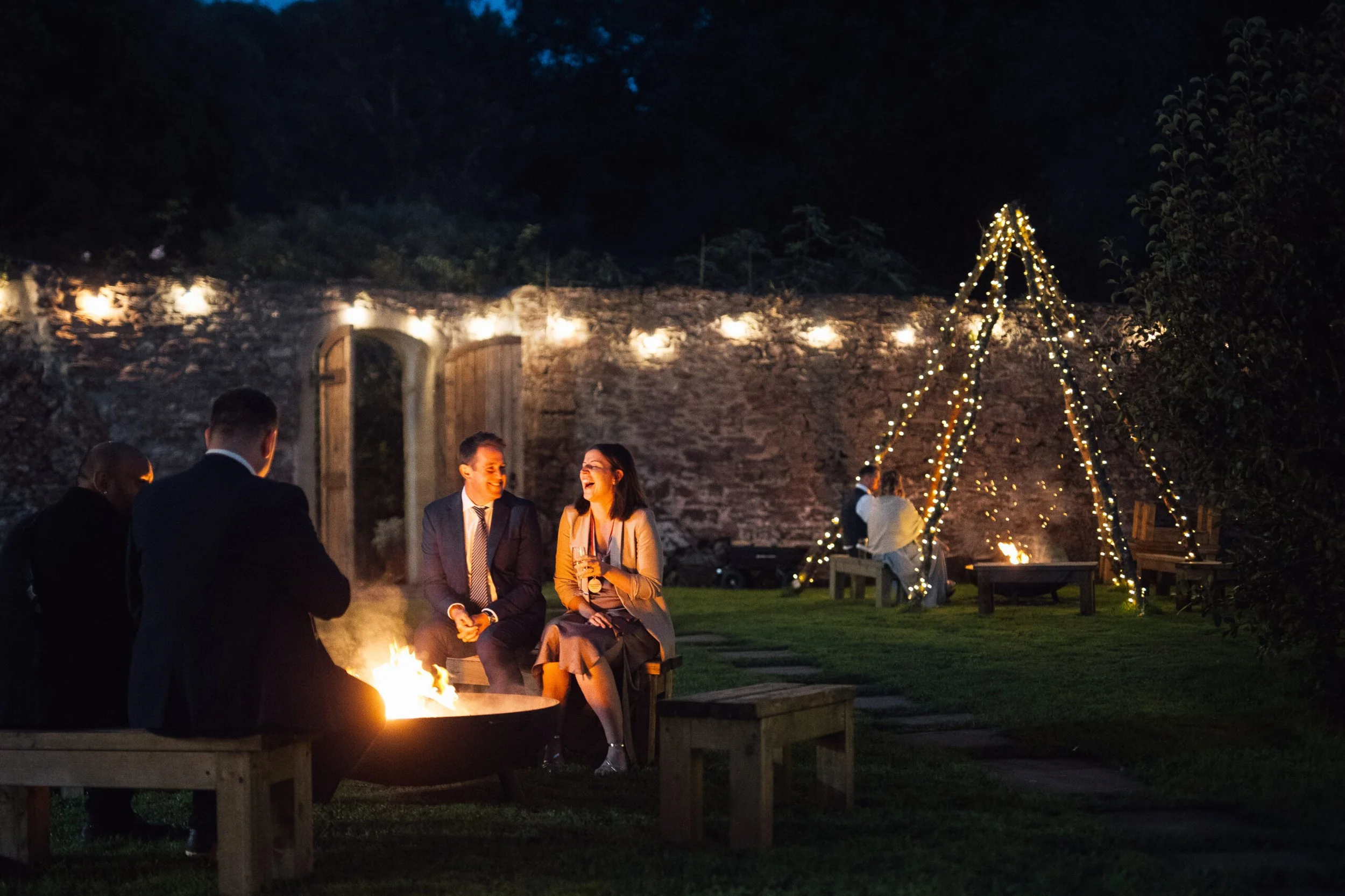 People gathered outdoors at night around a fire pit, engaging in conversation, with festive string lights and a decorated tree in the background.