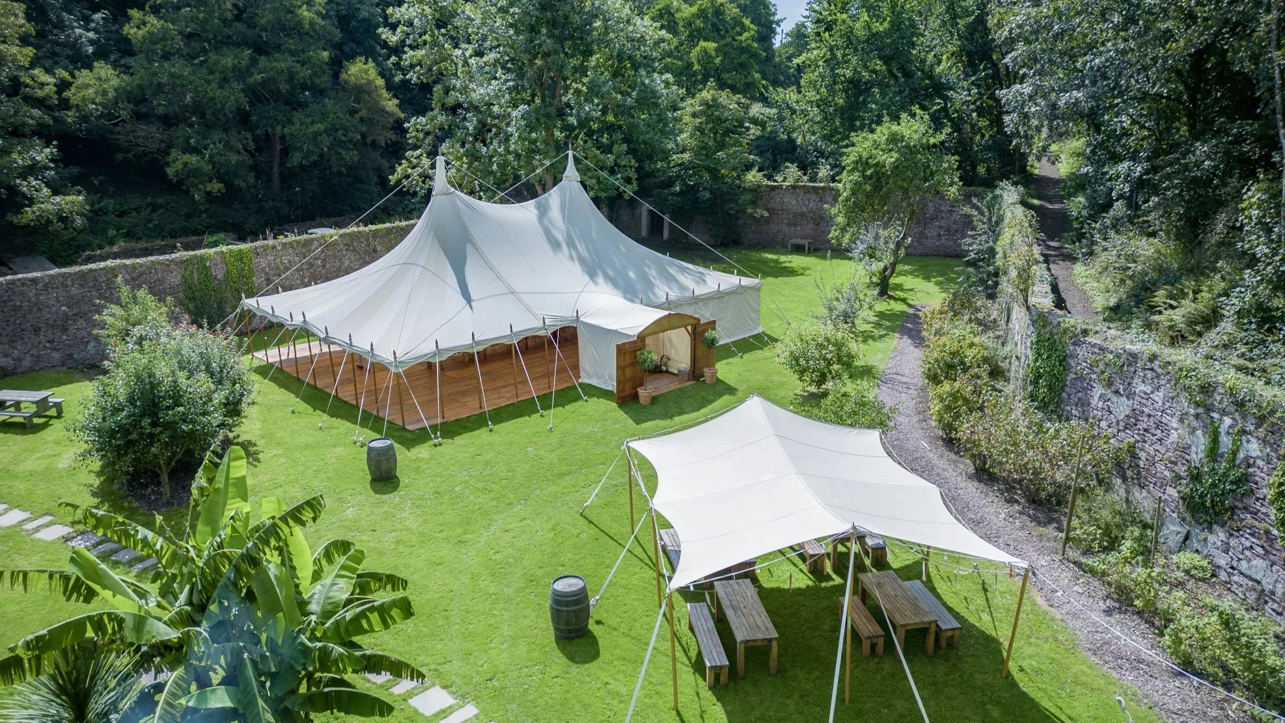 A large white tent set up in a manicured lawn surrounded by trees and stone walls, with additional smaller tents and outdoor furniture nearby.