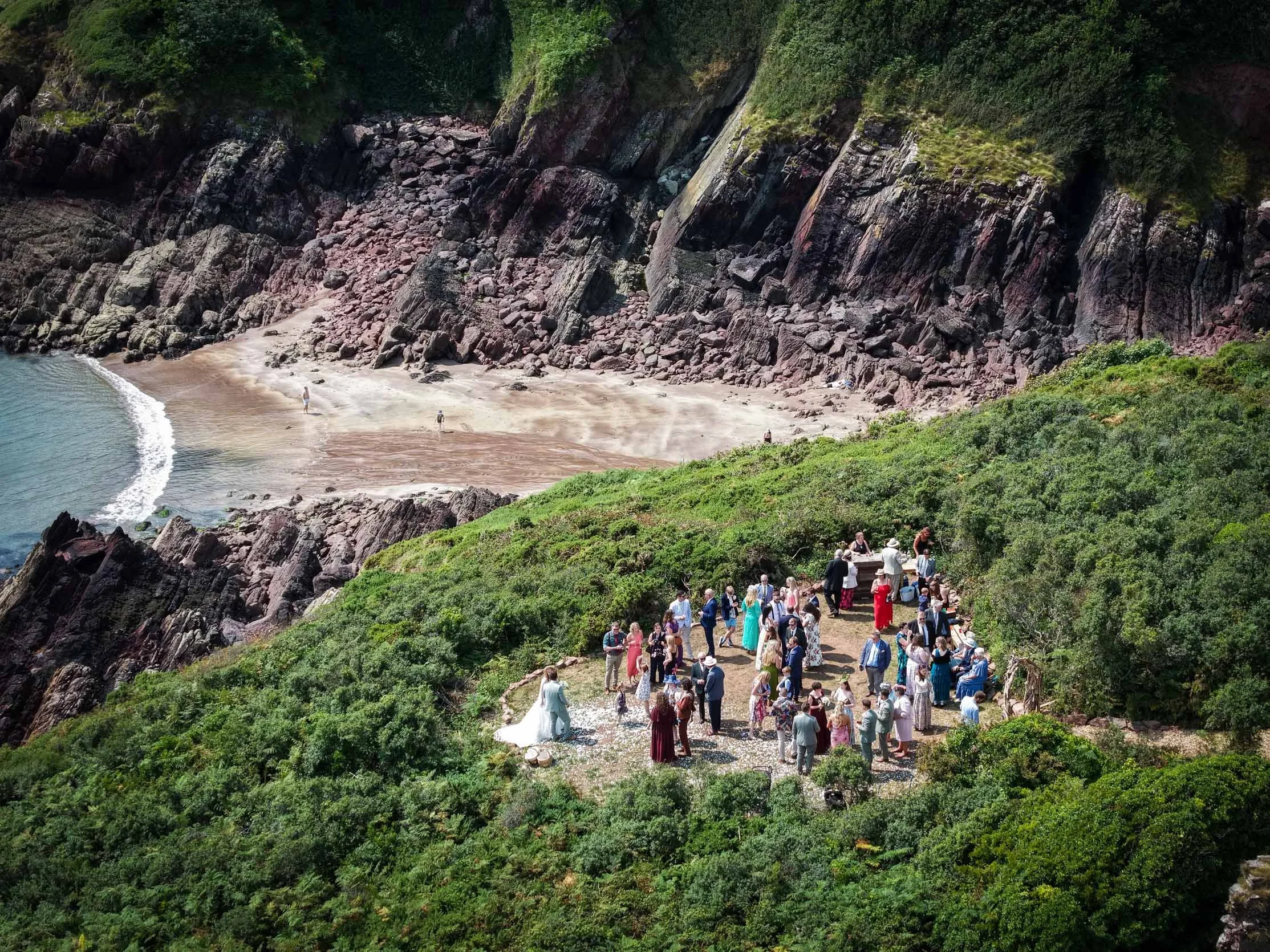 A large group of people dressed in formal attire gather on a path near cliffs and greenery, with a beach and water in the background.