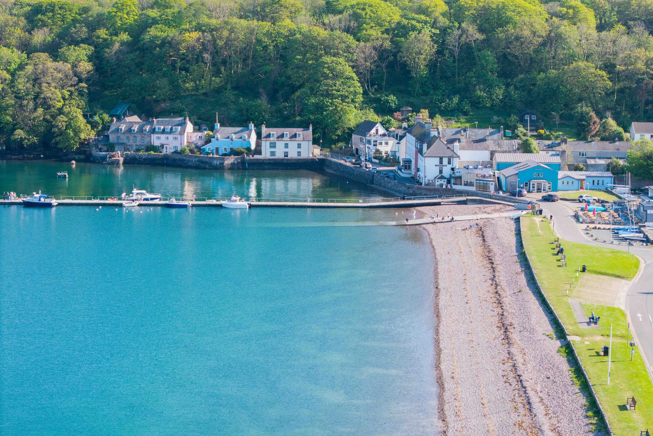 A coastal village with colorful houses along the water, boats docked at a pier, and green trees on a hillside in the background.