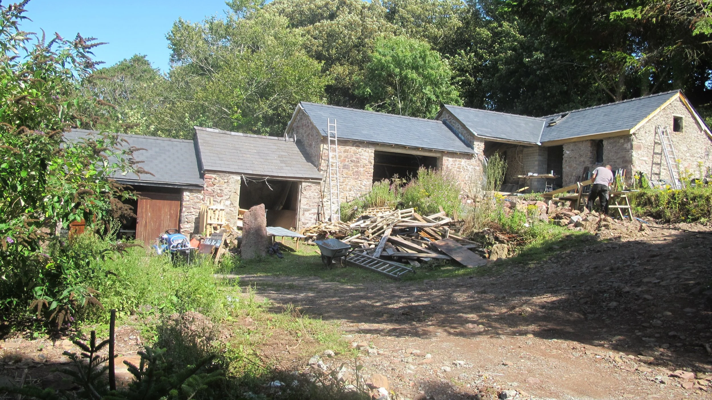 A stone house under renovation with construction tools, ladders, and a worker surrounded by trees and greenery.