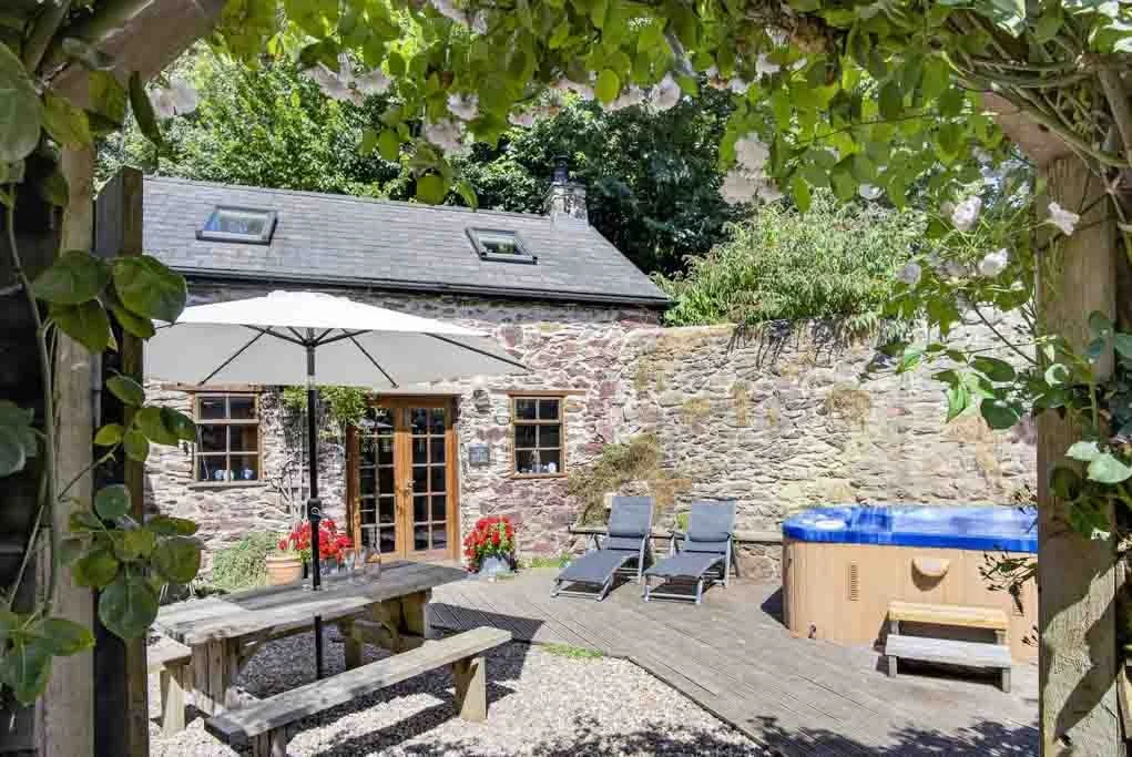 A cozy backyard patio scene showing a stone house with a slate roof, a white umbrella, a wooden picnic table with benches, two lounge chairs, and a hot tub, surrounded by lush greenery and flowering plants.