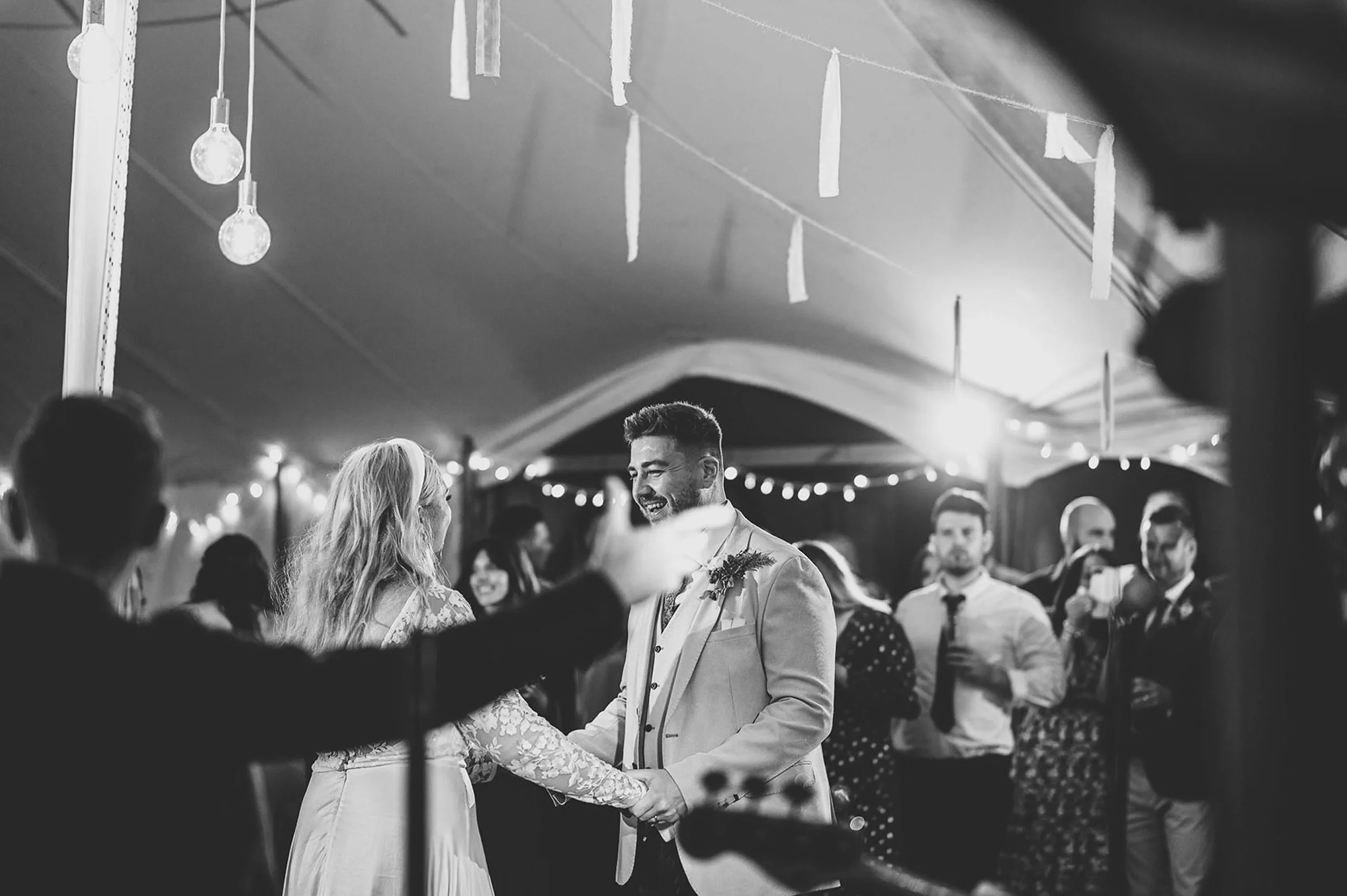 A couple in wedding attire dancing at their wedding reception, smiling, with guests and string lights in the background.