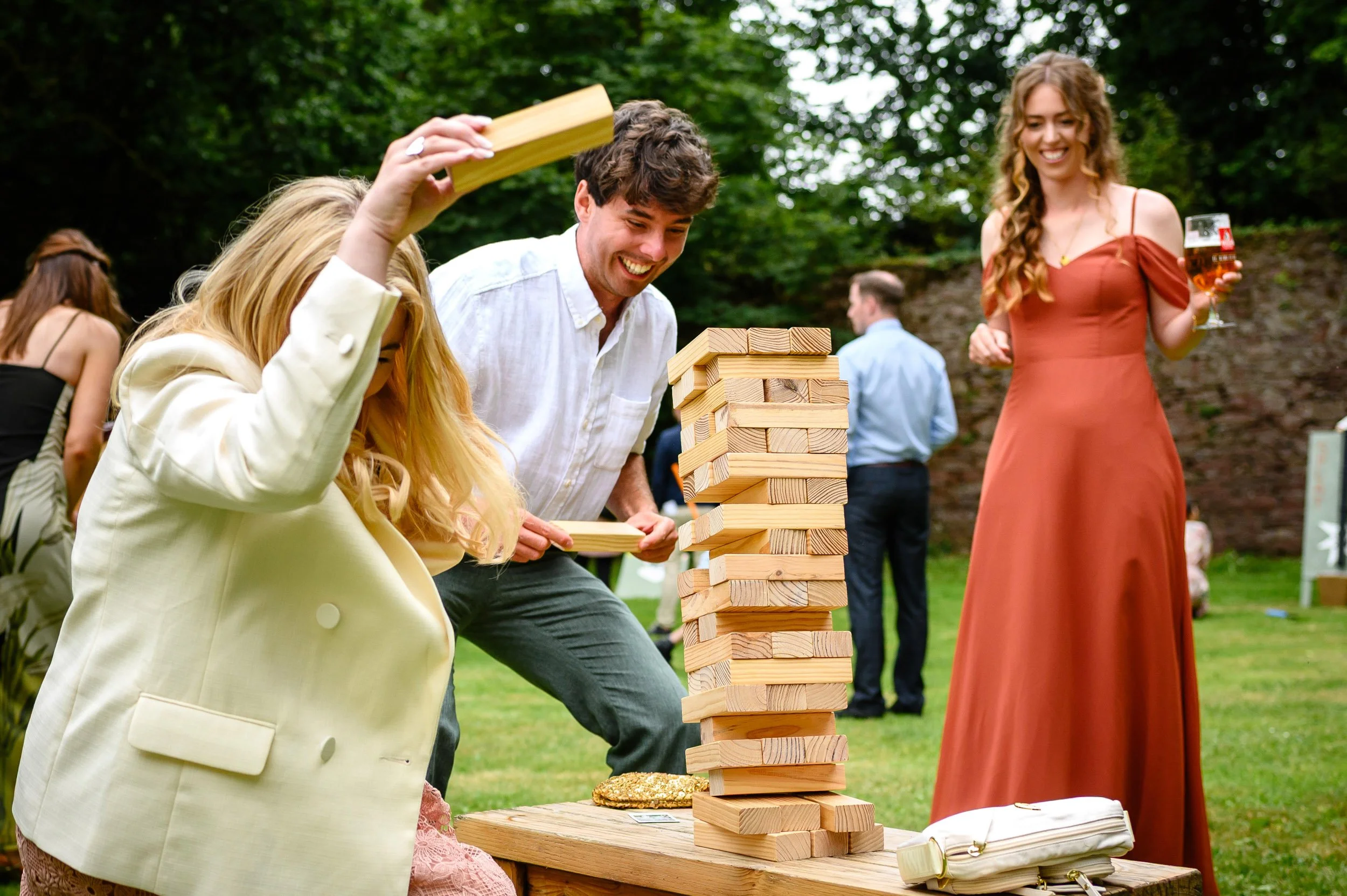 People playing giant Jenga outdoors at a garden party, with green trees in the background.