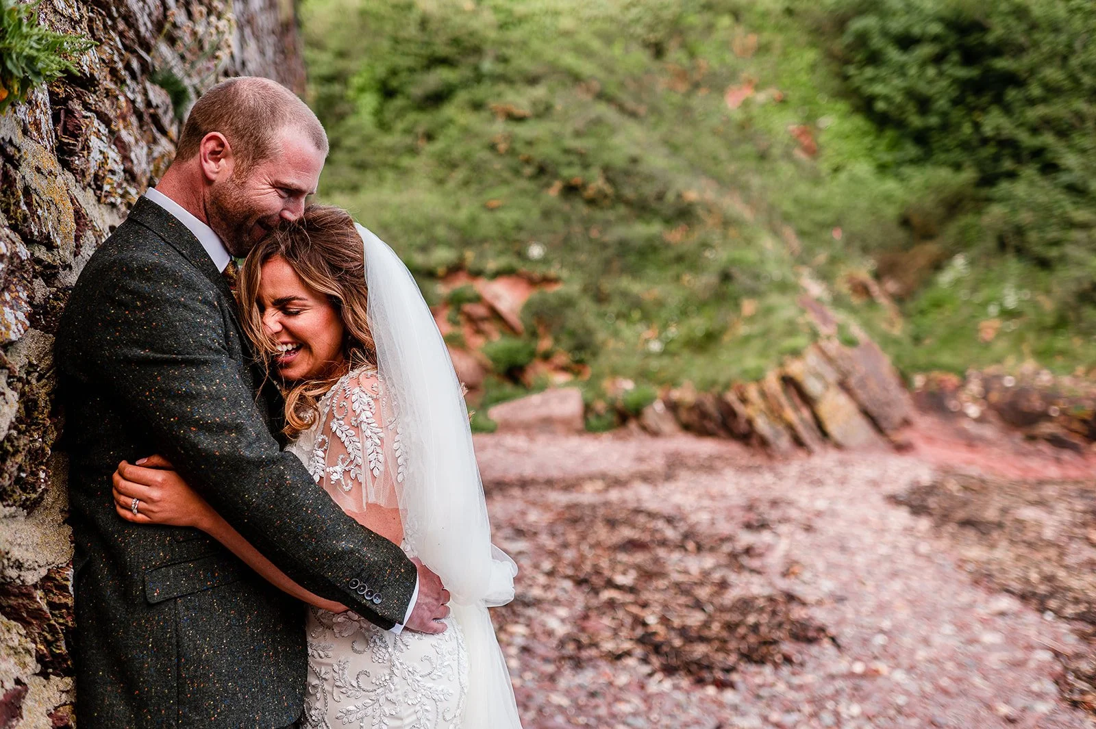 A newlywed couple is hugging and laughing together outside, leaning against a stone wall with a blurred natural background of mountains and trees.
