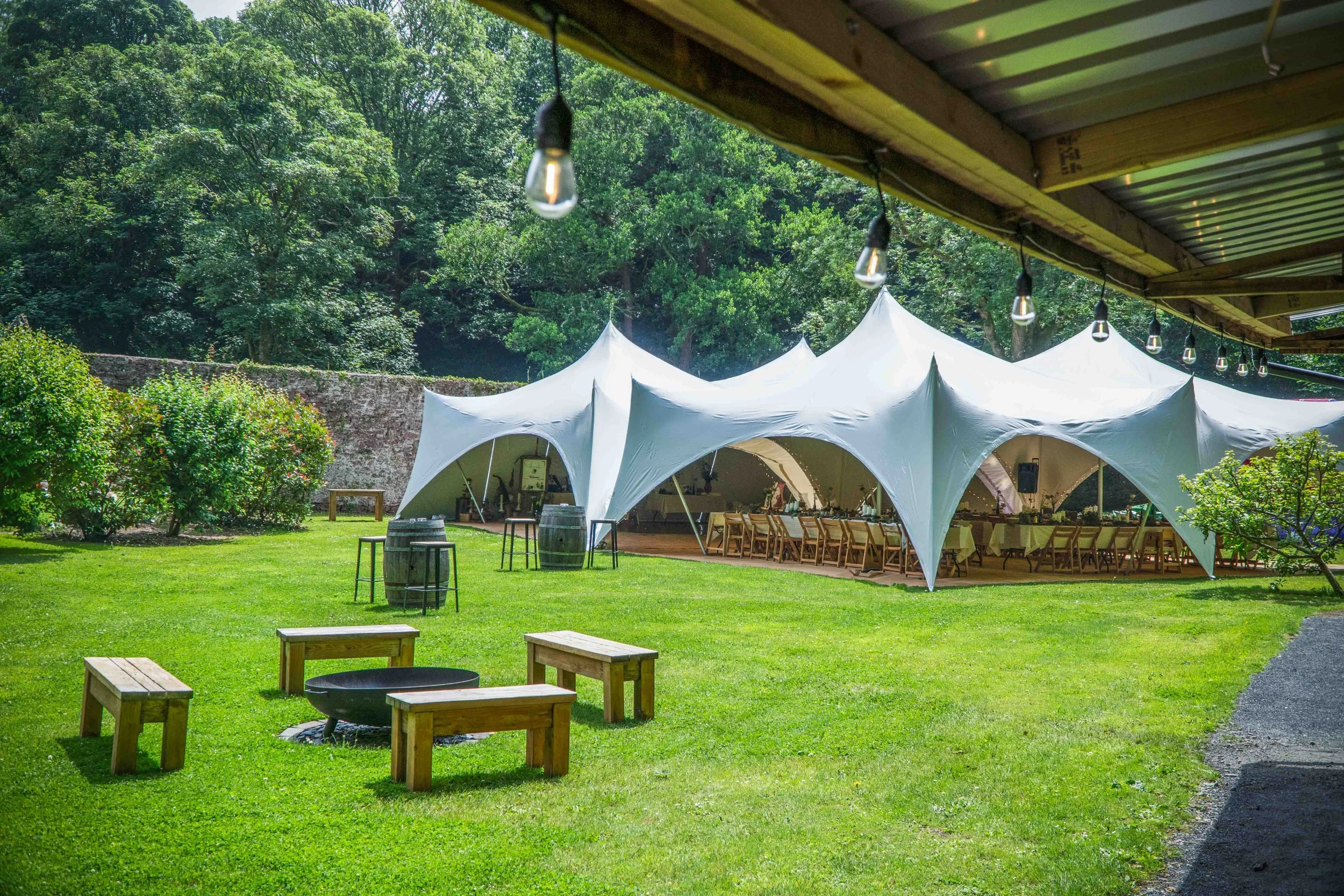 An outdoor event setup with white tents on a lush green lawn surrounded by trees, with string lights hanging from a wooden roof and wooden benches and barrels positioned around a firepit.