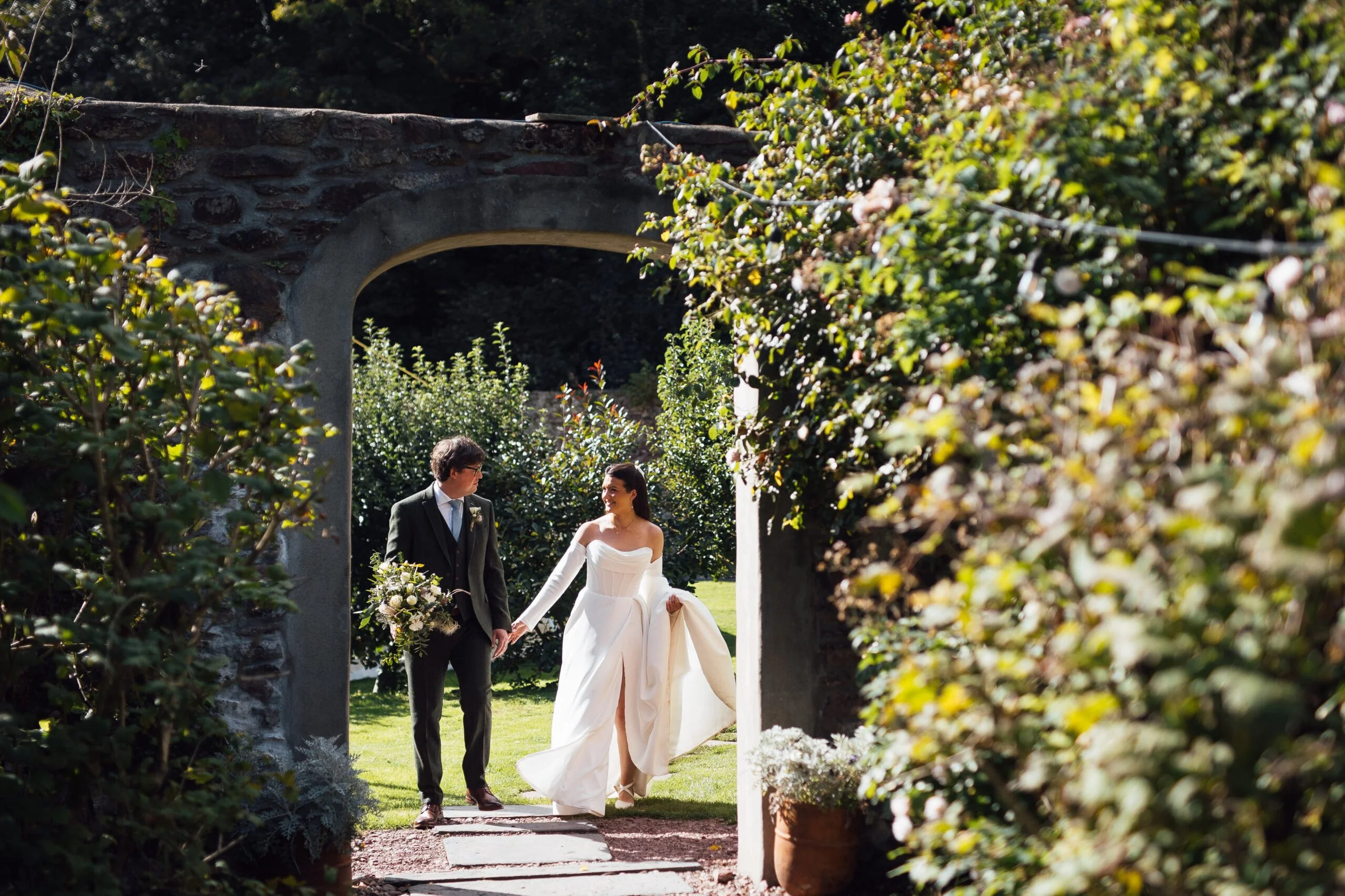 A bride and groom holding hands, walking through a garden archway decorated with lush greenery and flowers during an outdoor wedding ceremony.