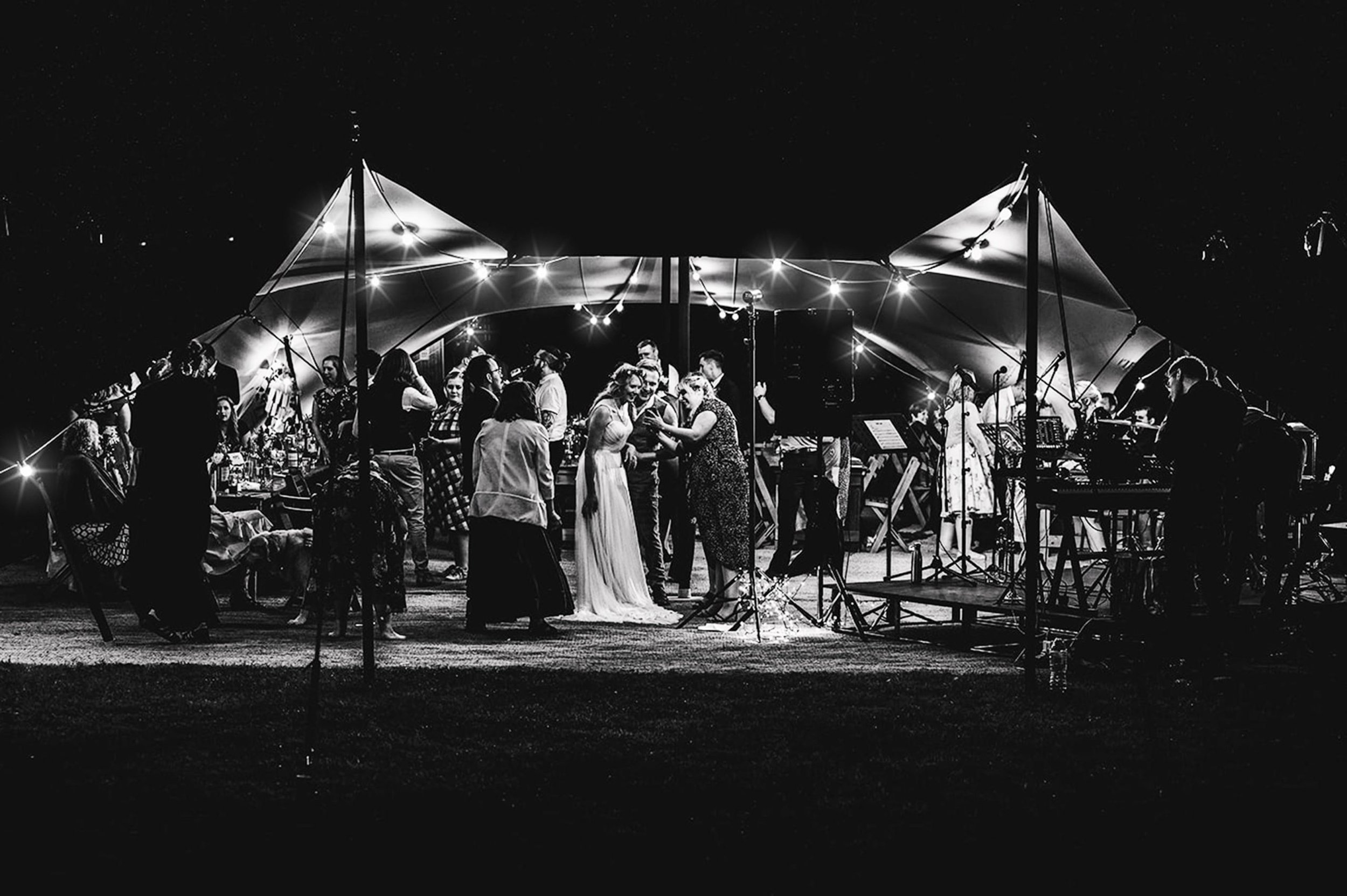 Black and white photo of a group of people dancing and socializing under a large outdoor canopy with string lights at night, near a stage with a band or musical setup.