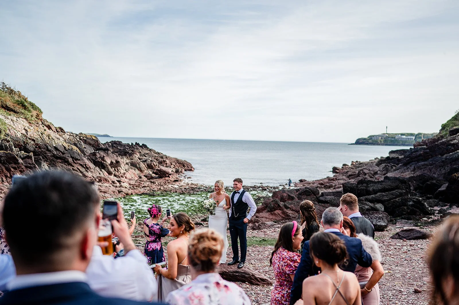 Wedding ceremony on a rocky beach with the bride and groom standing together, surrounded by guests, with the ocean and rocky cliffs in the background.