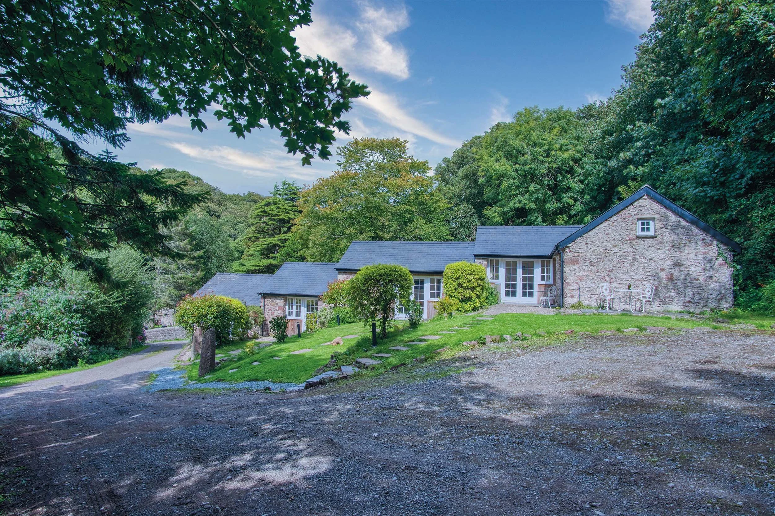 A scenic view of a stone house with a dark roof, surrounded by greenery and trees, with a clear blue sky overhead.