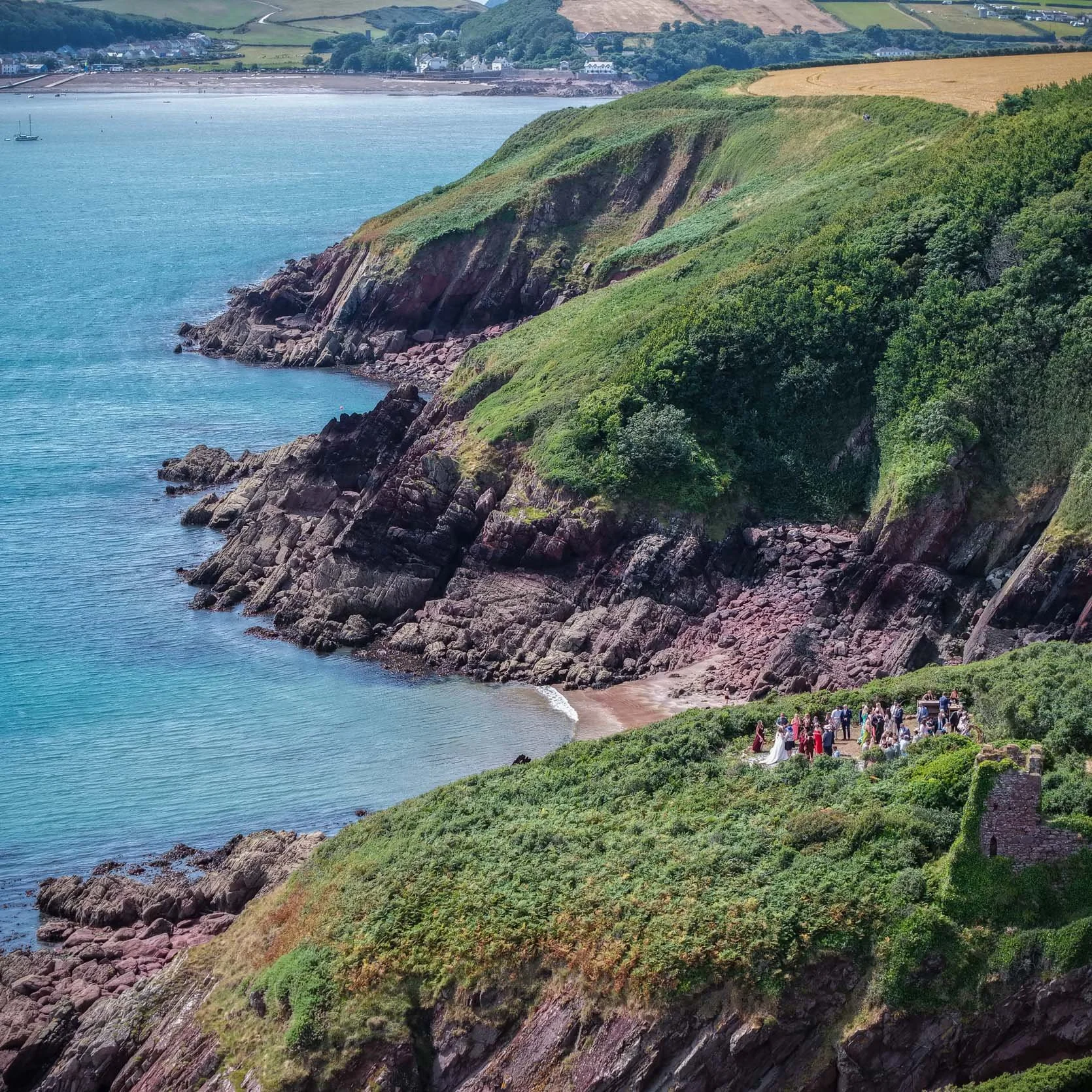 A group of people gathered on a grassy hillside overlooking the ocean with rocky cliffs. In the background, there are houses and fields on rolling hills.