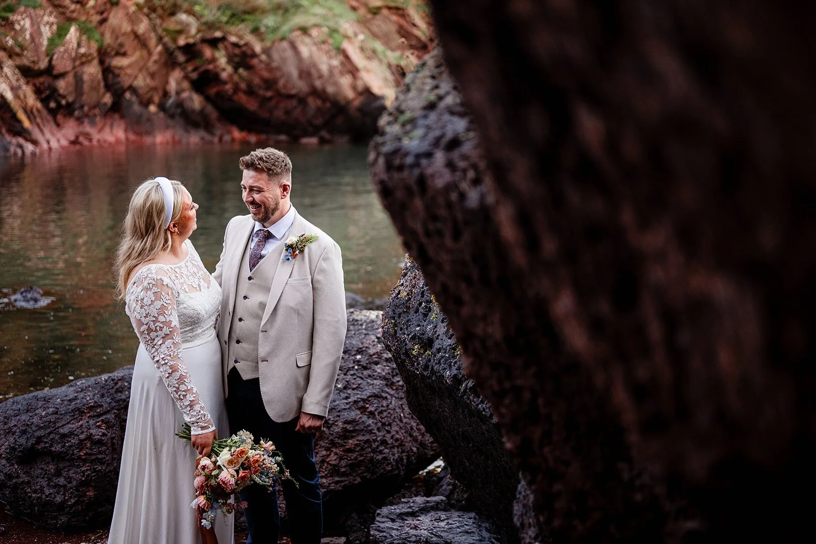 A bride and groom in wedding attire standing by a river with rocky banks, smiling at each other, with the groom holding a bouquet of flowers.