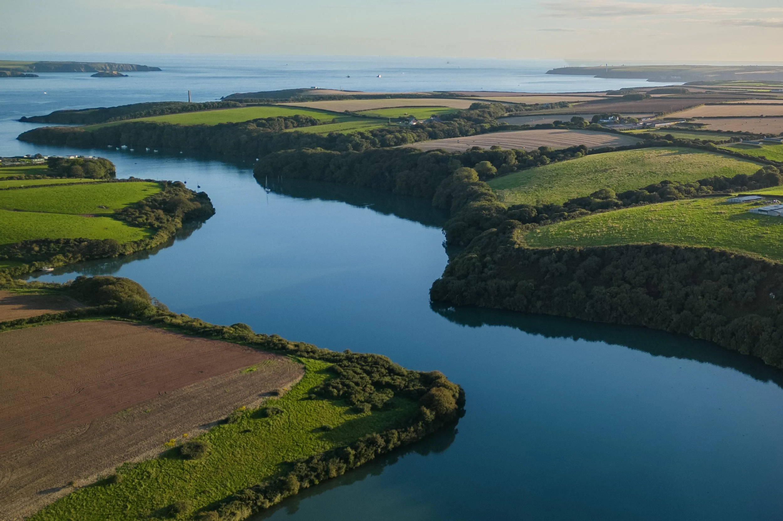 Aerial view of green fields, a river, and the ocean in the distance with islands and boats, under a partly cloudy sky.