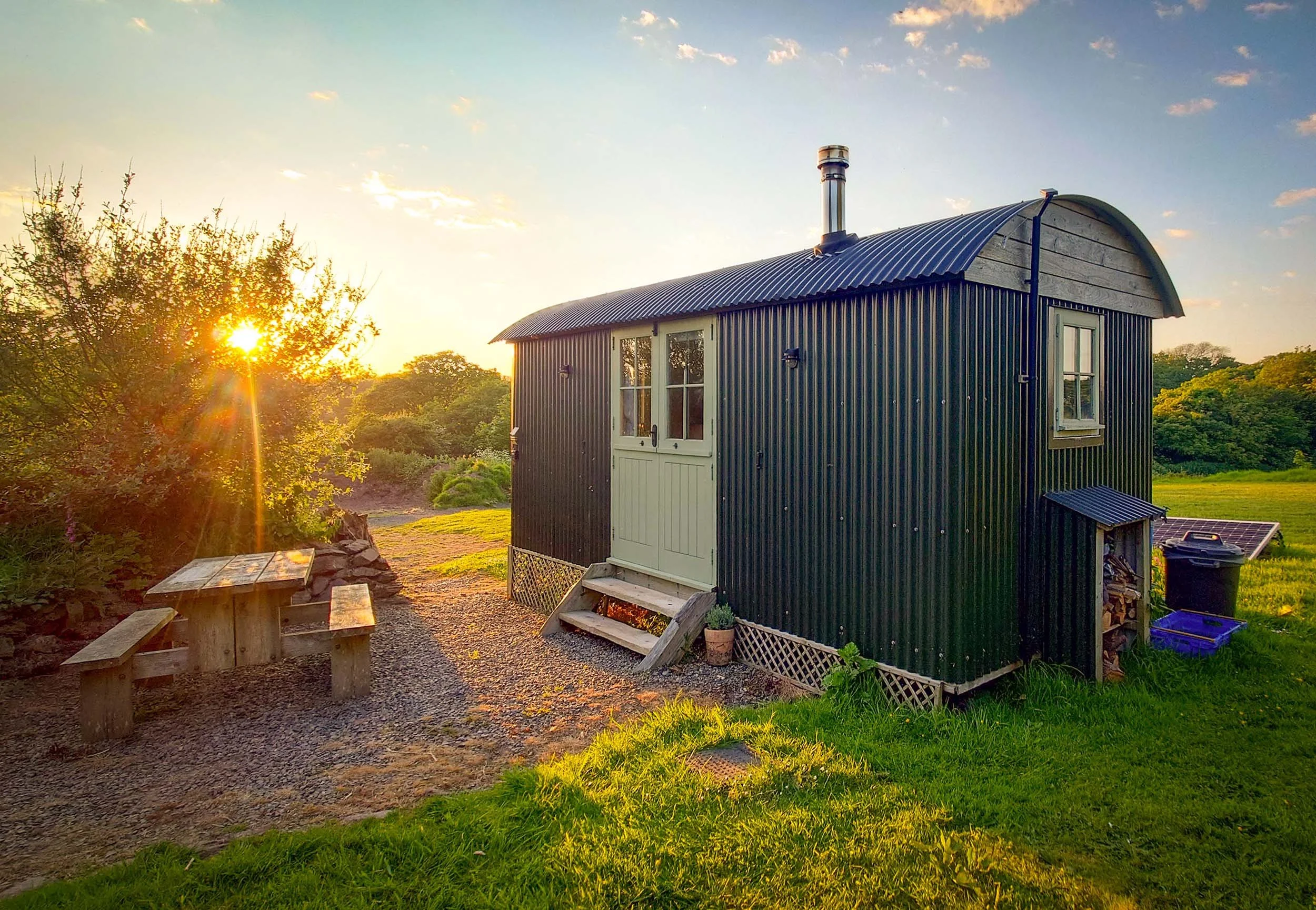 A small black metal building with white double doors and windows, set in a scenic outdoor area during sunset with trees and green grass, and a woodpile underneath a small roof extension.