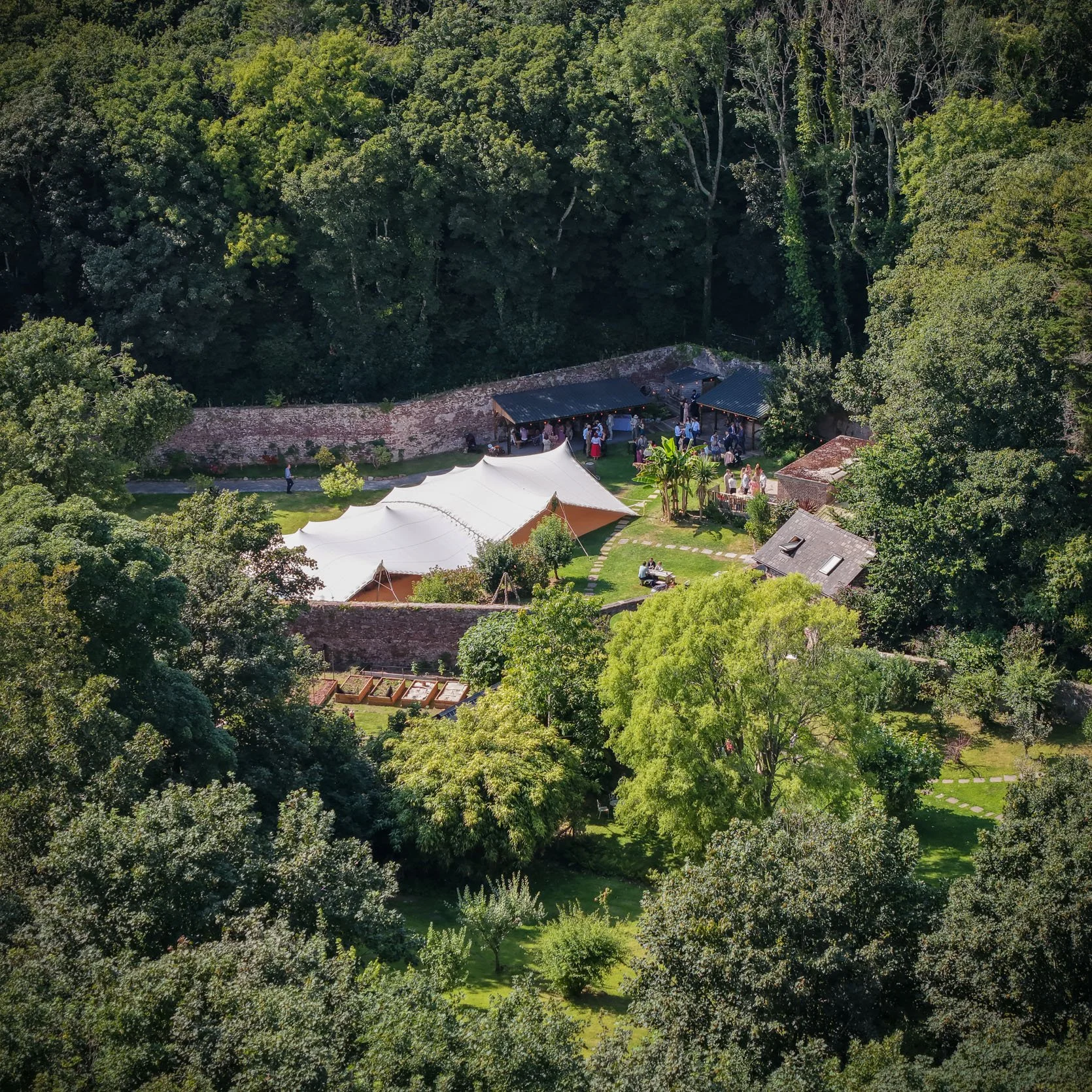 Aerial view of a garden during a social event, with a large white event tent, smaller structures, and groups of people gathered among the trees and around the tents.