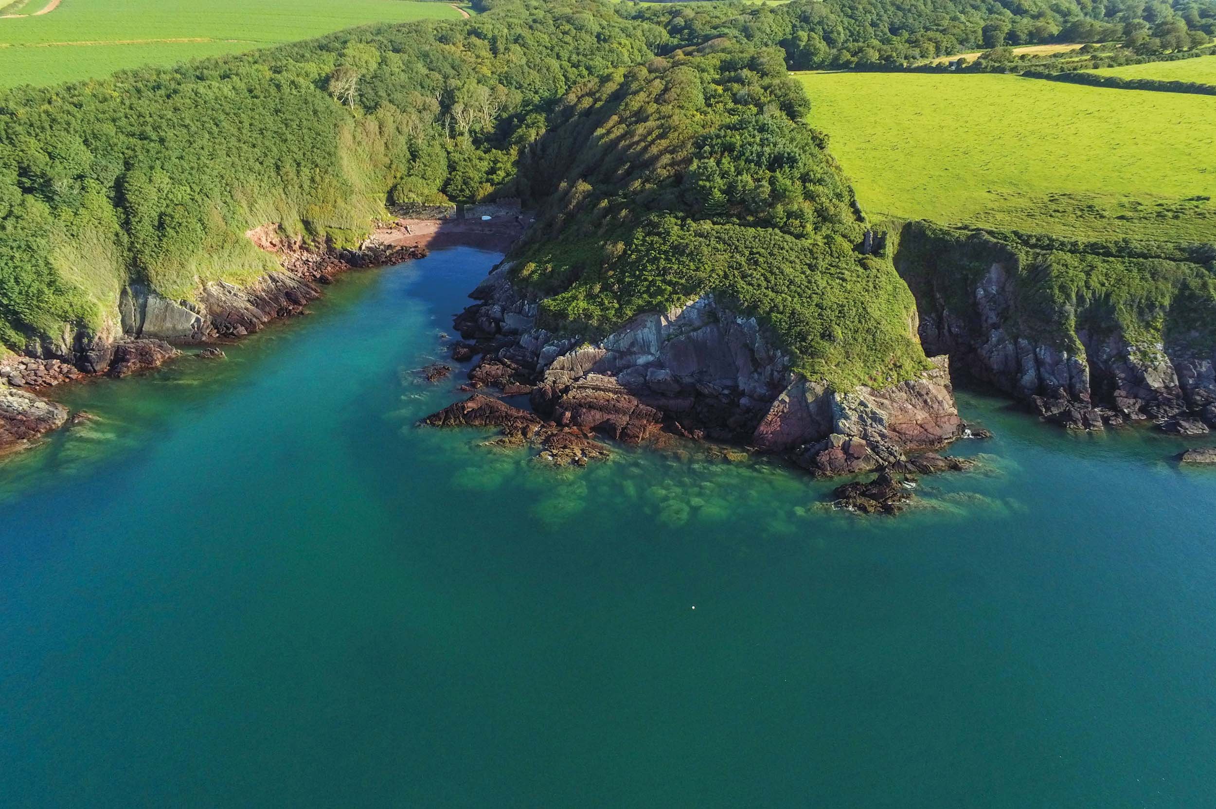 Aerial view of a narrow cove with turquoise water, surrounded by lush green hills and rocky shorelines.