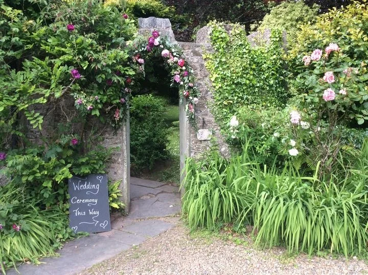 Stone garden archway decorated with pink and white flowers, leading into a lush green garden with a wooden pathway, and a black signboard reading 'Wedding Ceremony This Way' on the left side.