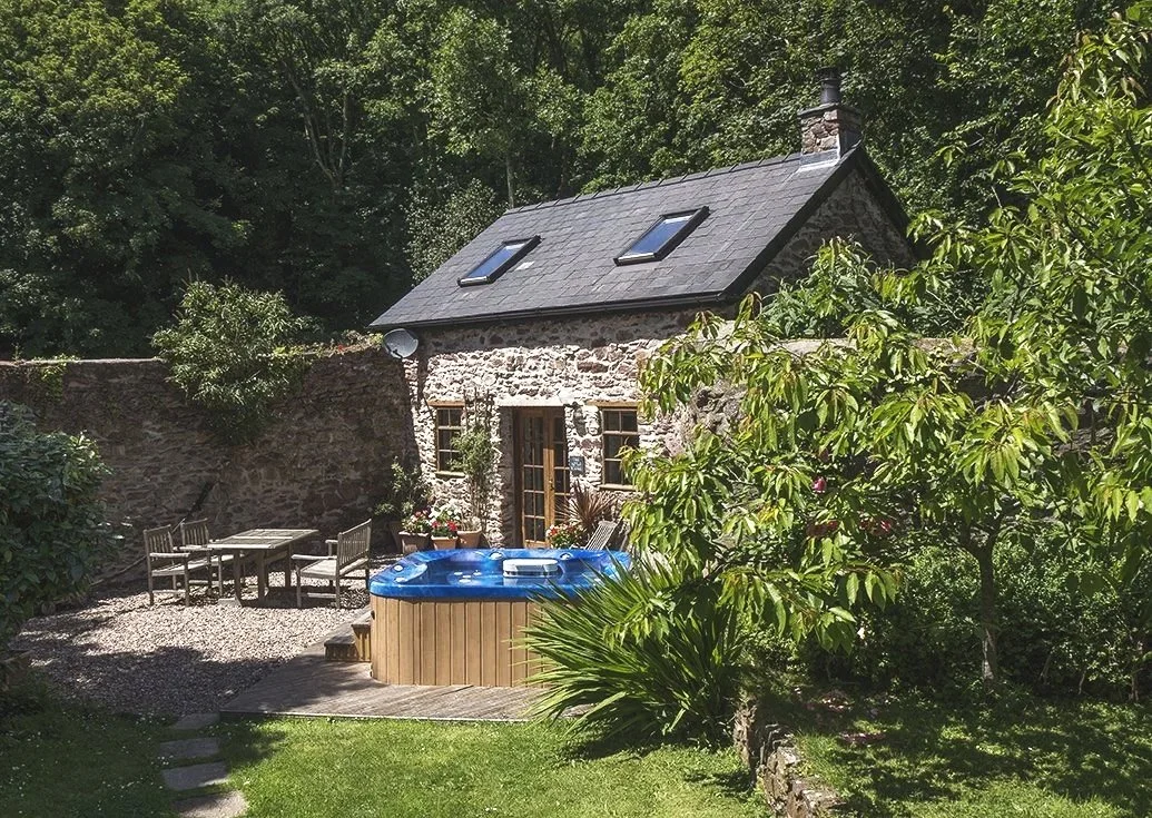 A small stone cottage surrounded by green trees and plants, with a deck featuring an outdoor table and chairs, and a hot tub in the backyard.