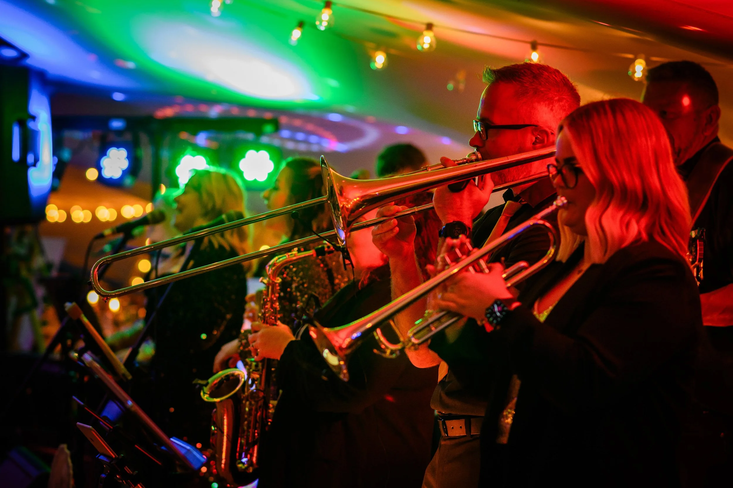 A group of musicians playing trombones and other instruments in a vibrant, colourful wedding reception, illuminated by multi-coloured stage lights and string lights.
