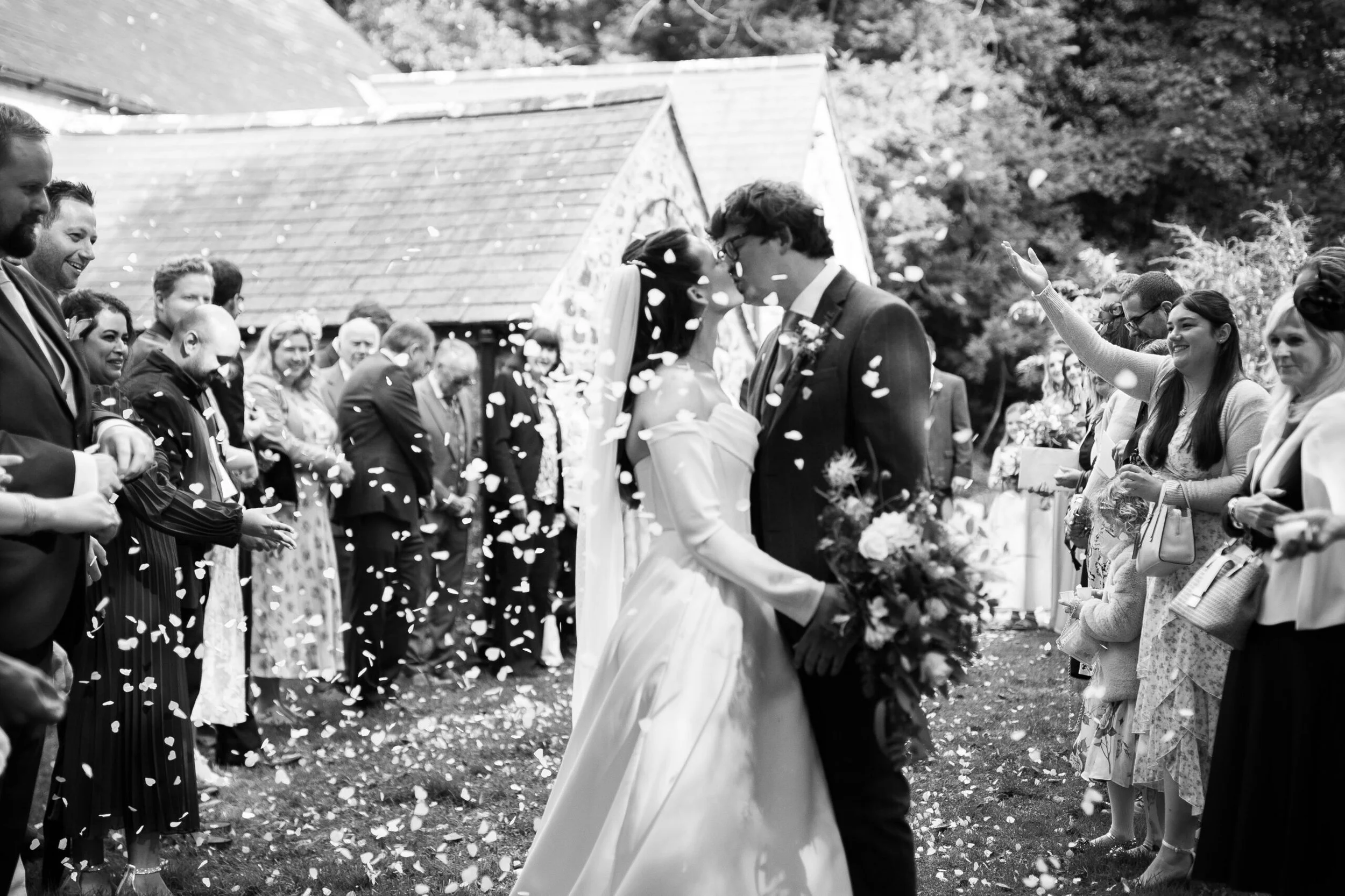 A black and white photo of a wedding scene with a bride and groom kissing, surrounded by guests throwing flower petals.