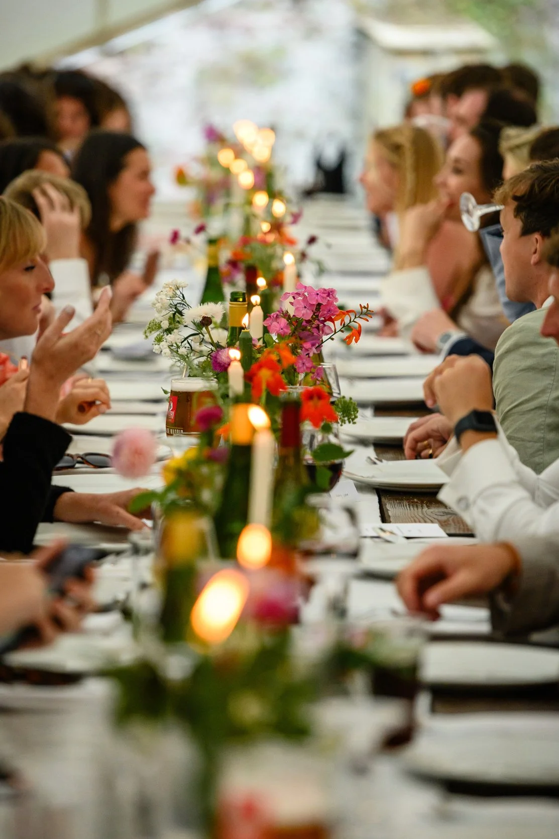 A long banquet table with people dining and conversing, decorated with colorful flowers and lit candles, in a bright setting.