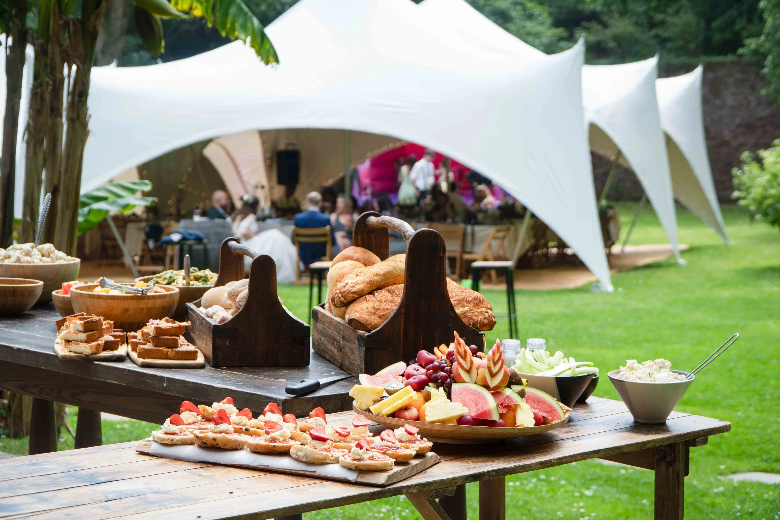 A buffet table set outdoors with various foods including breads, fruits, and desserts, under a large white canopy with people dining in the background.