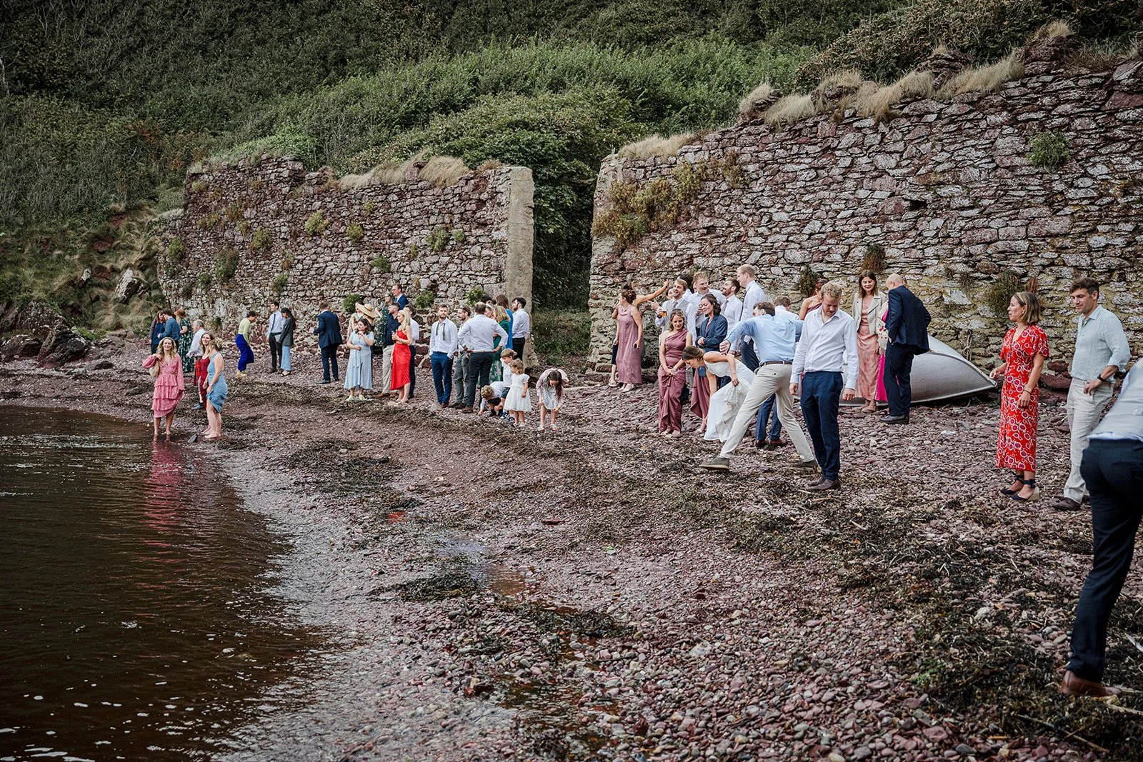 A group of people in formal and semi-formal attire gathered on a rocky shoreline, some wading in the water, near an old stone wall and lush greenery.