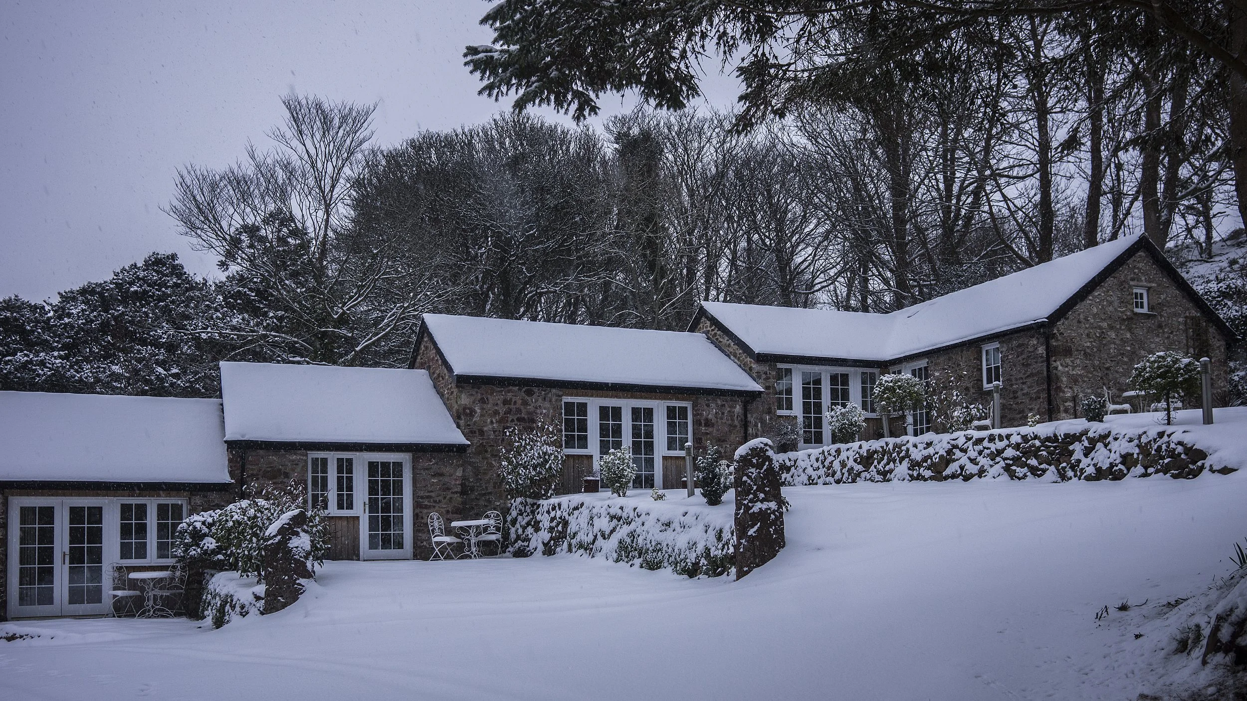 Snow-covered rustic stone house with multiple rooflines and white-framed windows, surrounded by snow-laden trees and a stone wall.