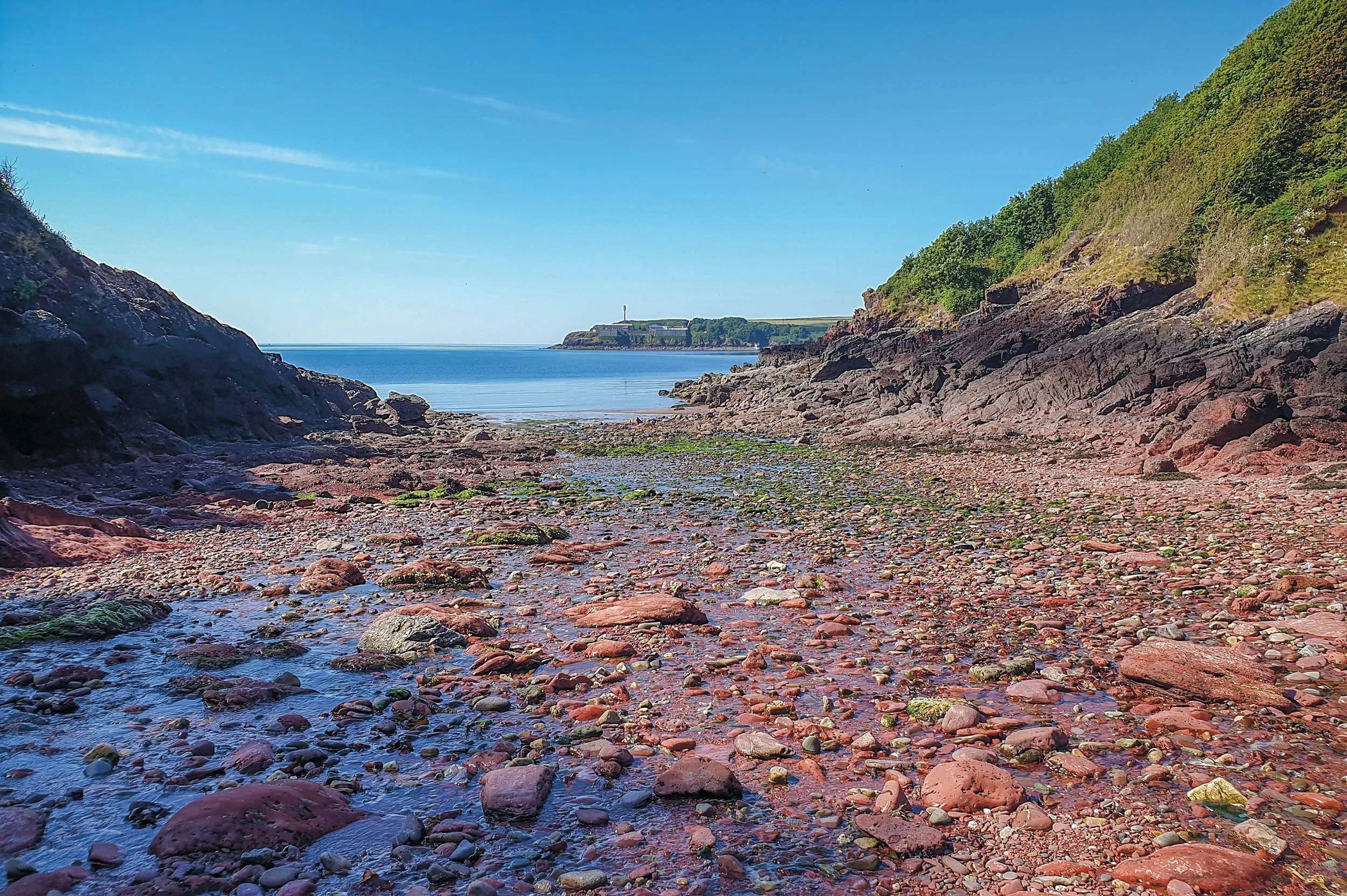 A rocky seaside cove with calm blue water, green hills on either side, and a building with a chimney in the distance.