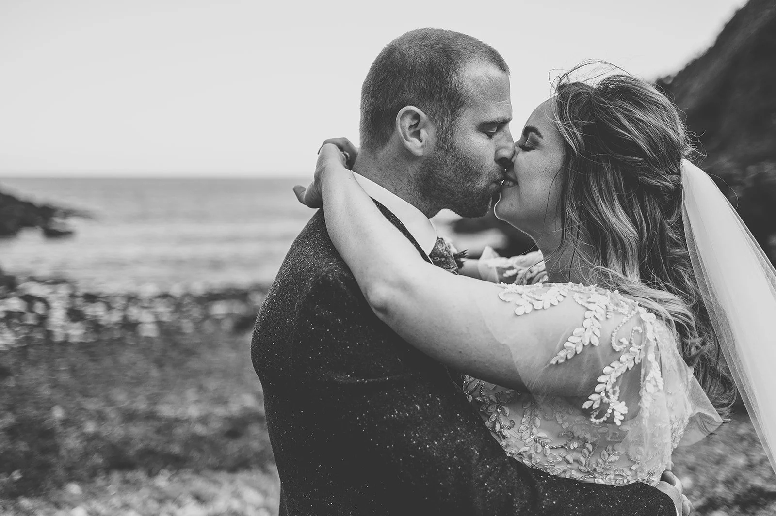 Black and white photo of a bride and groom kissing outdoors by the water, with rocky shoreline and cliffs in background.