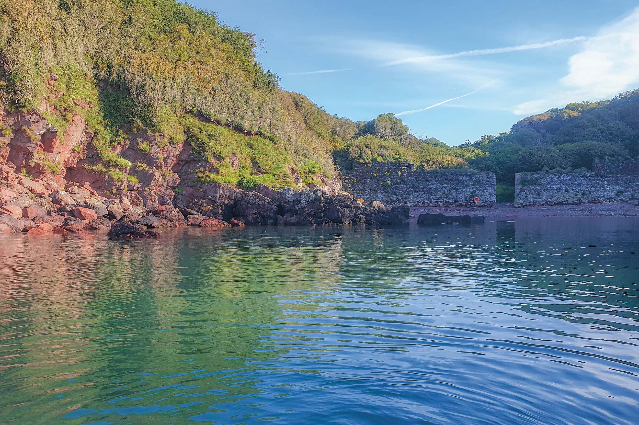 A body of water with gentle ripples, bordered by rocky shores and lush green trees on a hillside, under a clear blue sky with some clouds.