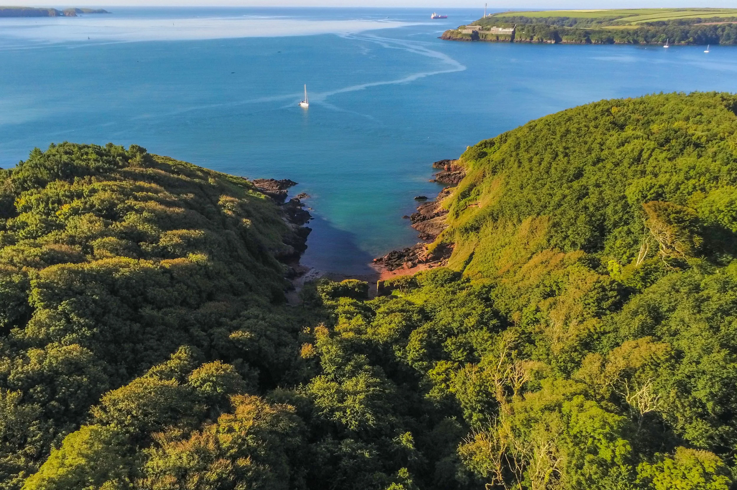 Aerial view of a coastal landscape with green hills, rocky shoreline, calm blue water, sailboats, and a distant cargo ship.