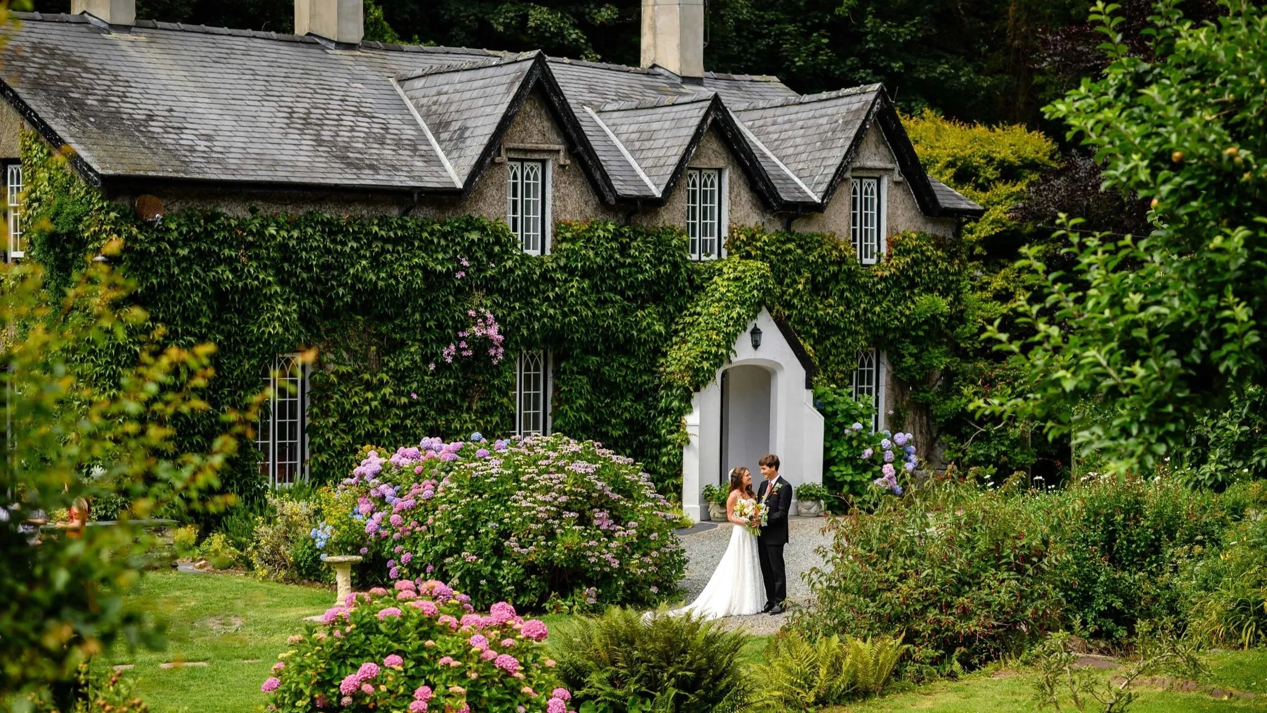 A bride and groom standing outside a stone house with ivy-covered walls, surrounded by colorful flowers and greenery.